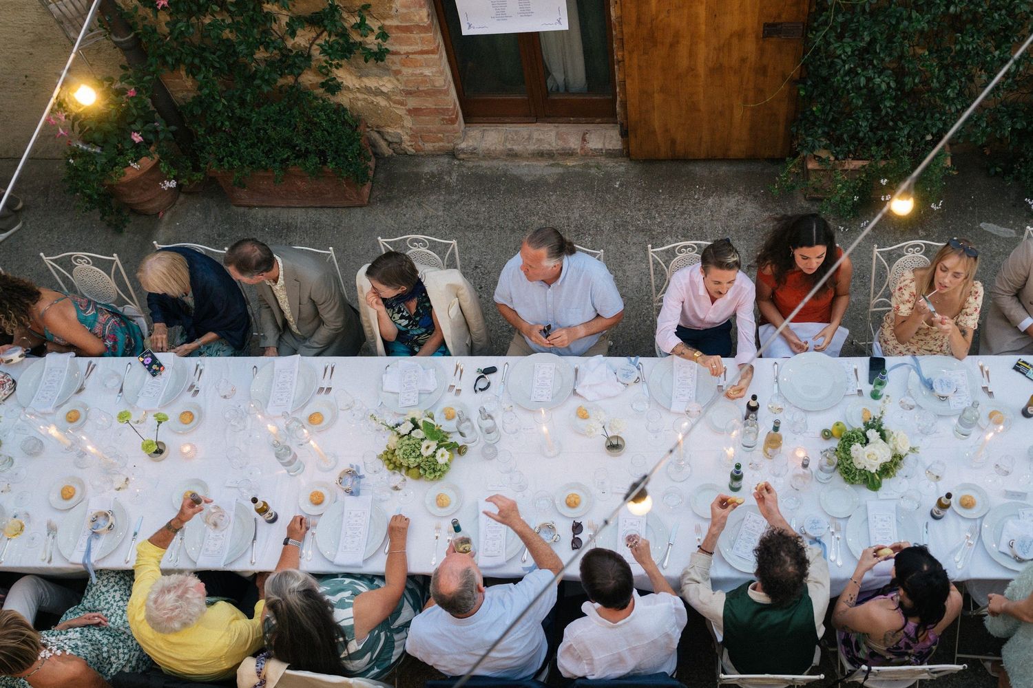 Aerial view of wedding guests toasting at long decorated table under string lights at outdoor reception.