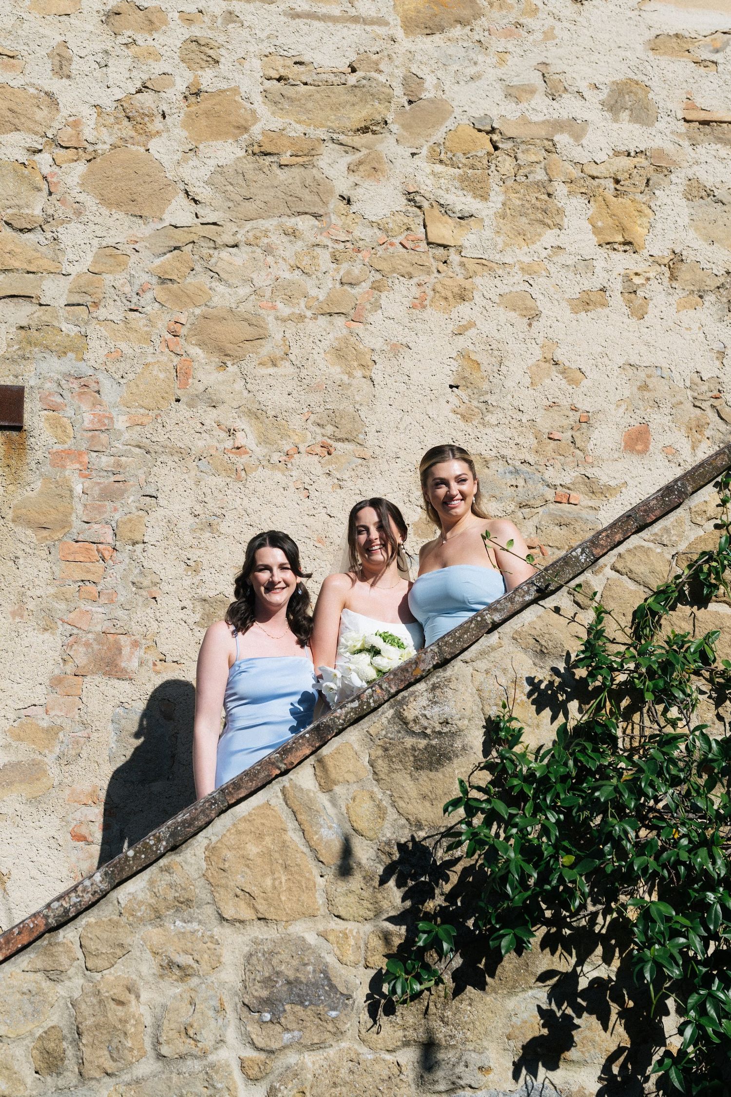 Three bridesmaids in light blue dresses posing on stone staircase against rustic wall.