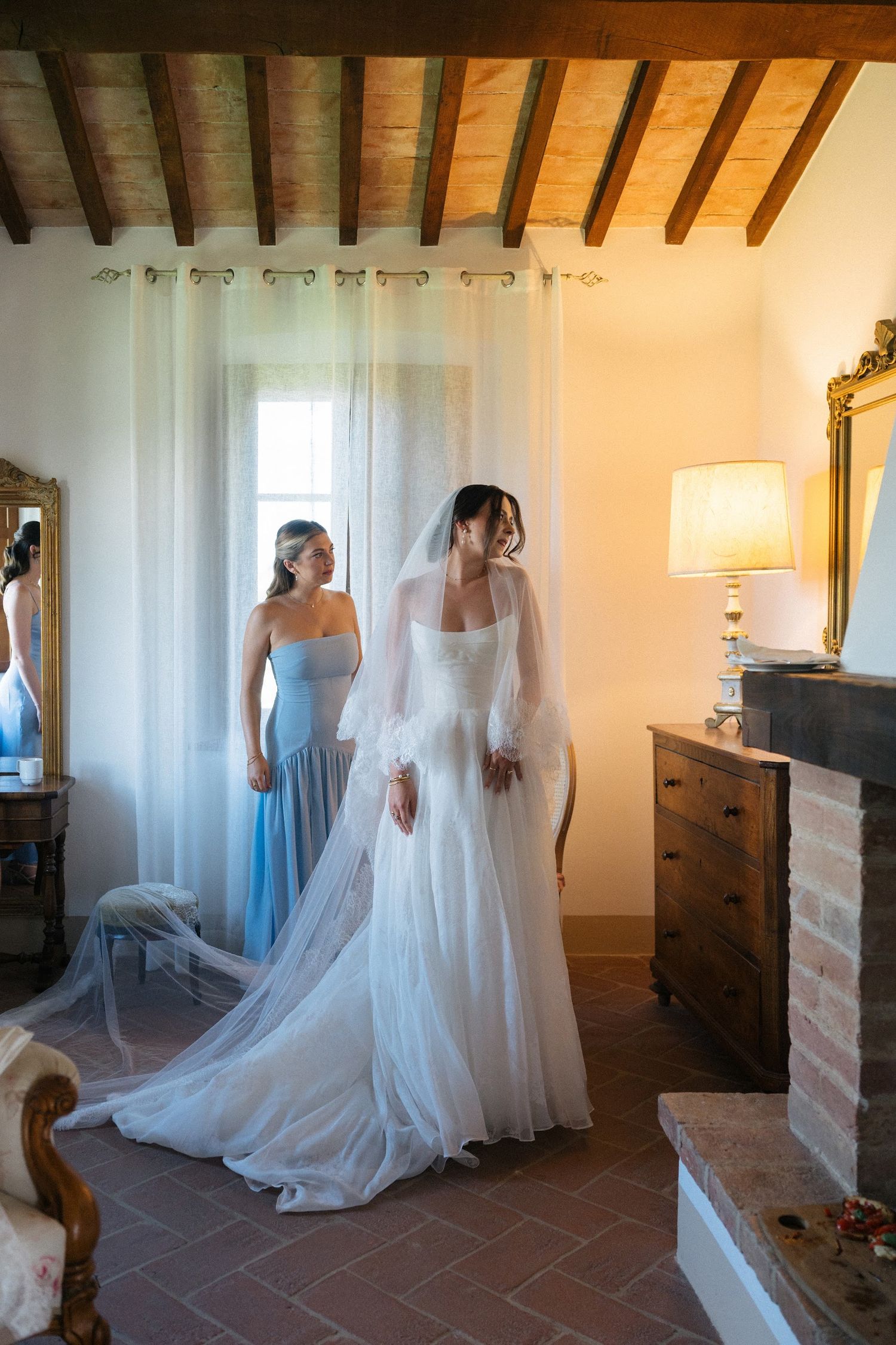 Bride in flowing white wedding gown with veil standing in rustic Tuscan villa room with wood beam ceiling.