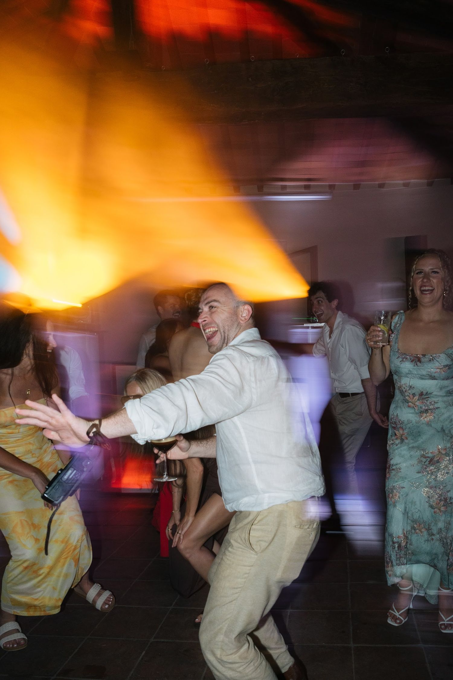 Joyful bald wedding guest dances energetically under dramatic orange lights at reception.