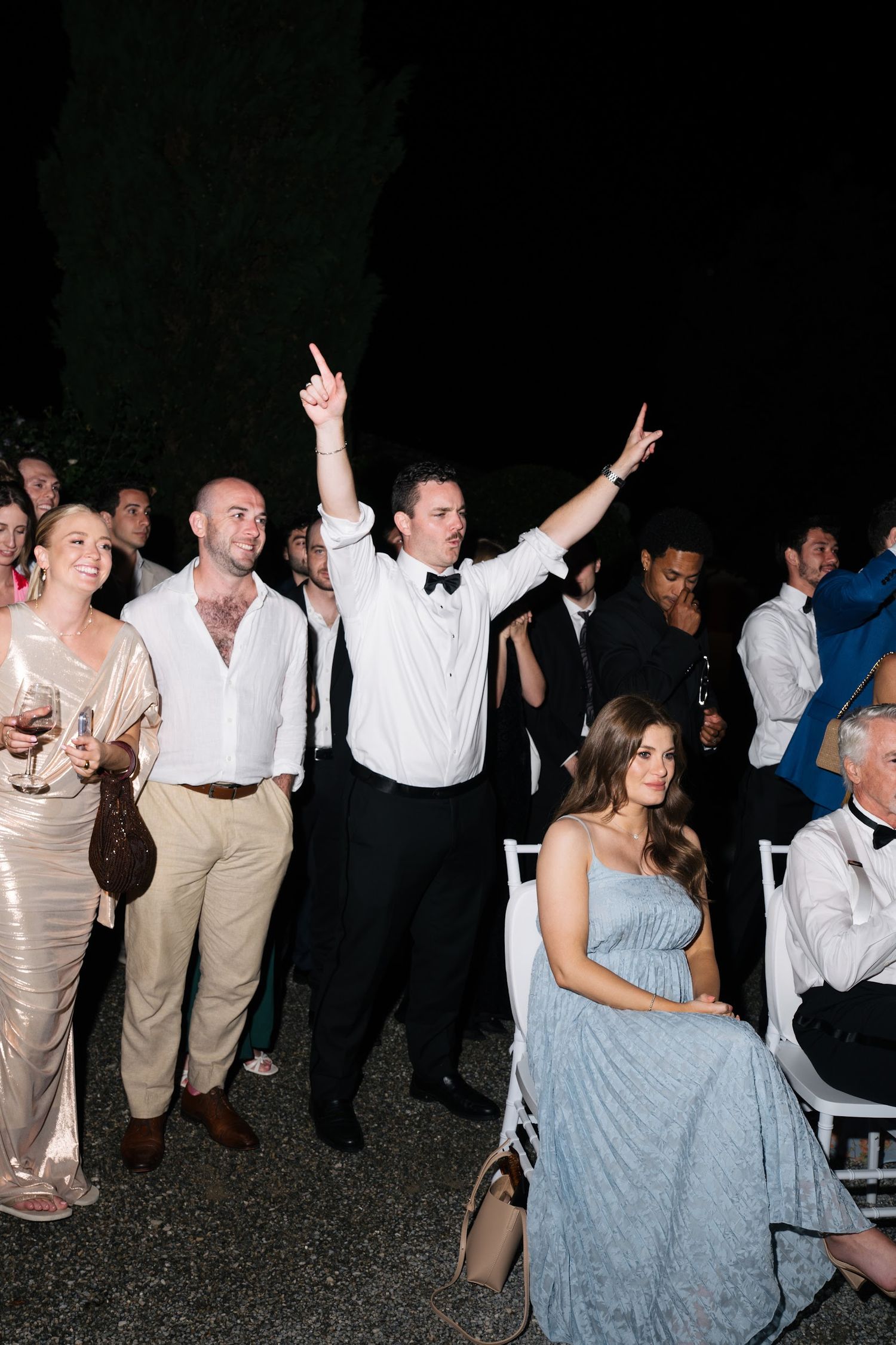 Excited wedding guest celebrating with hands raised among a crowd at an outdoor evening reception.