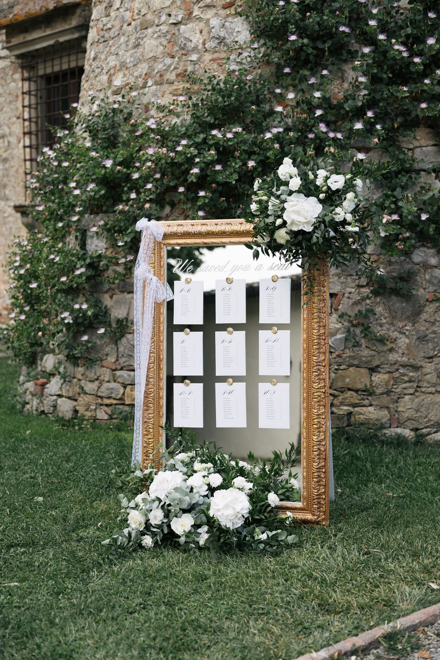 Elegant gold-framed mirror seating chart decorated with white roses and greenery at outdoor wedding.