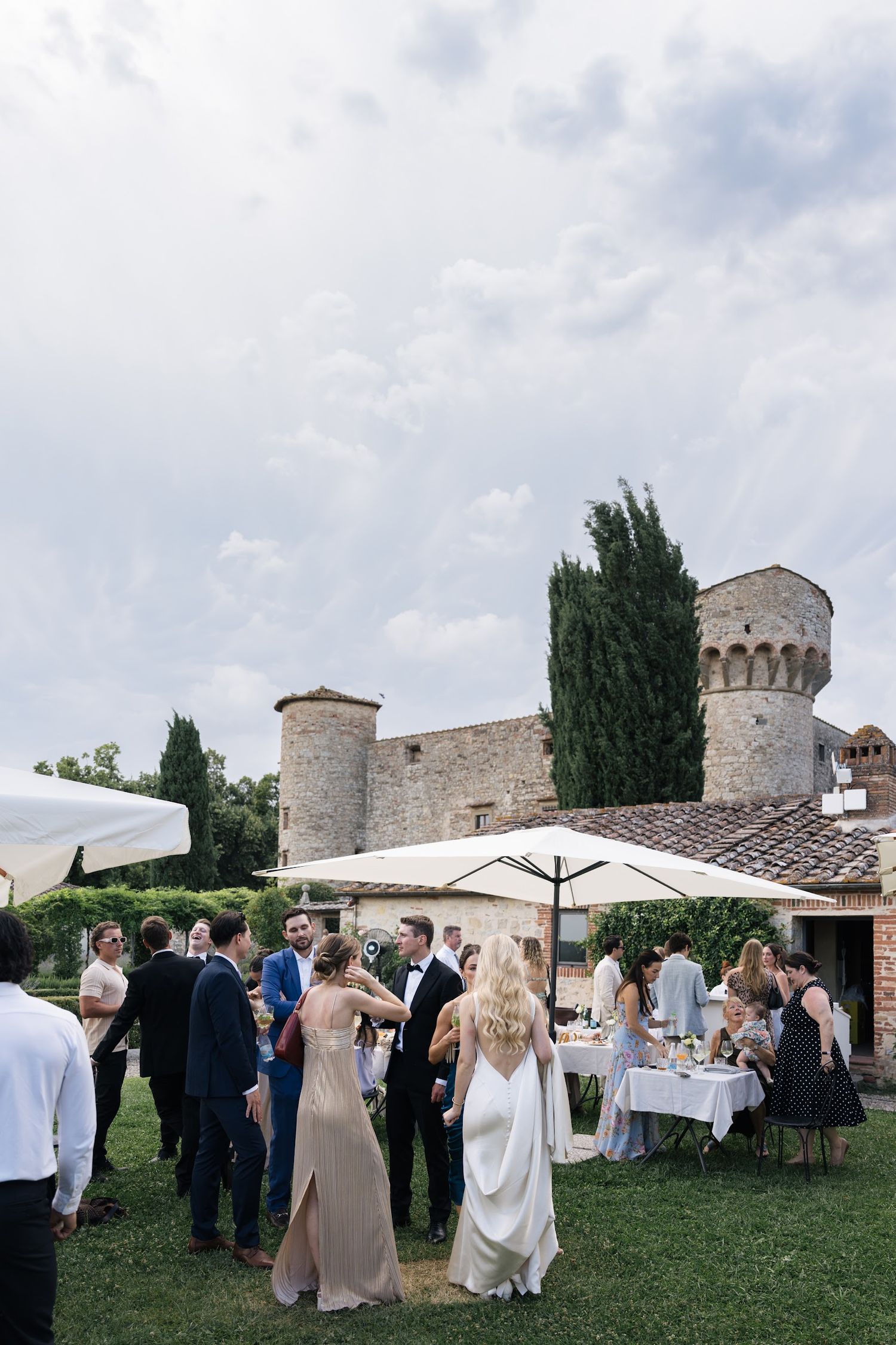 Wedding guests mingling on a lawn beside a medieval stone castle tower in Tuscany, Italy.