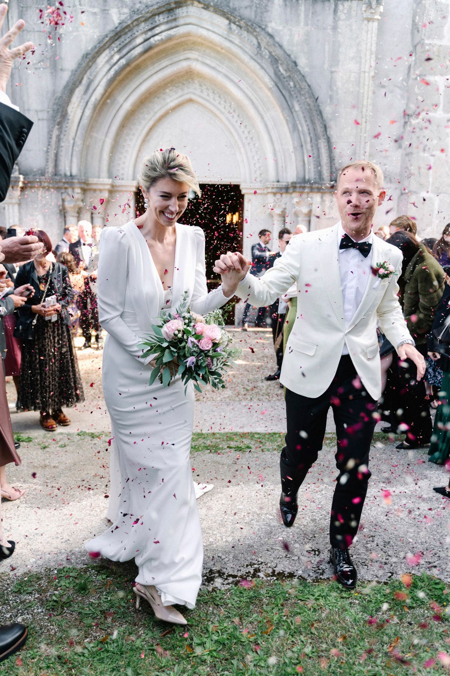 Joyful newlyweds exit church amid colorful confetti shower, bride in white gown, groom in white tuxedo jacket.