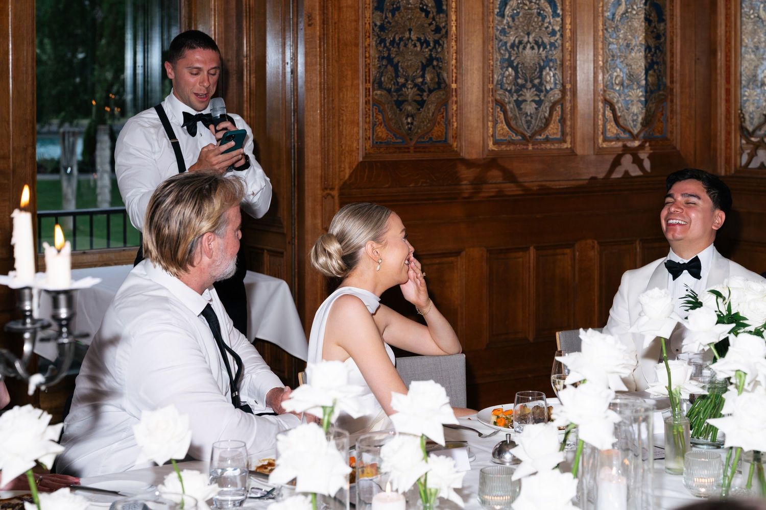 Bride and groom laugh together at wedding reception table while photographer captures the moment.