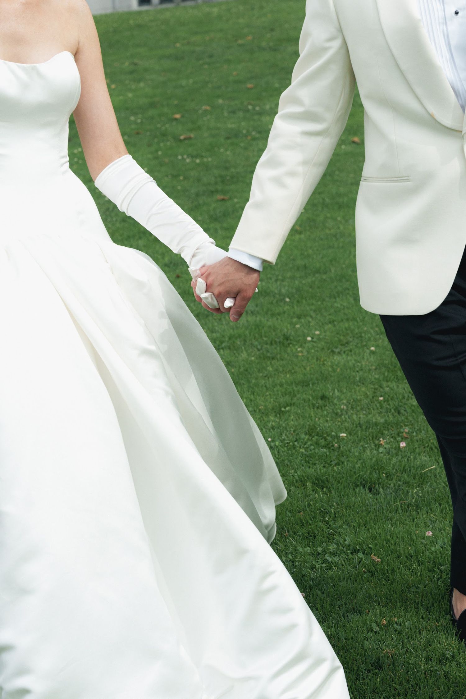 Bride and groom holding hands on green grass, bride in white strapless gown with gloves, groom in white tuxedo jacket.