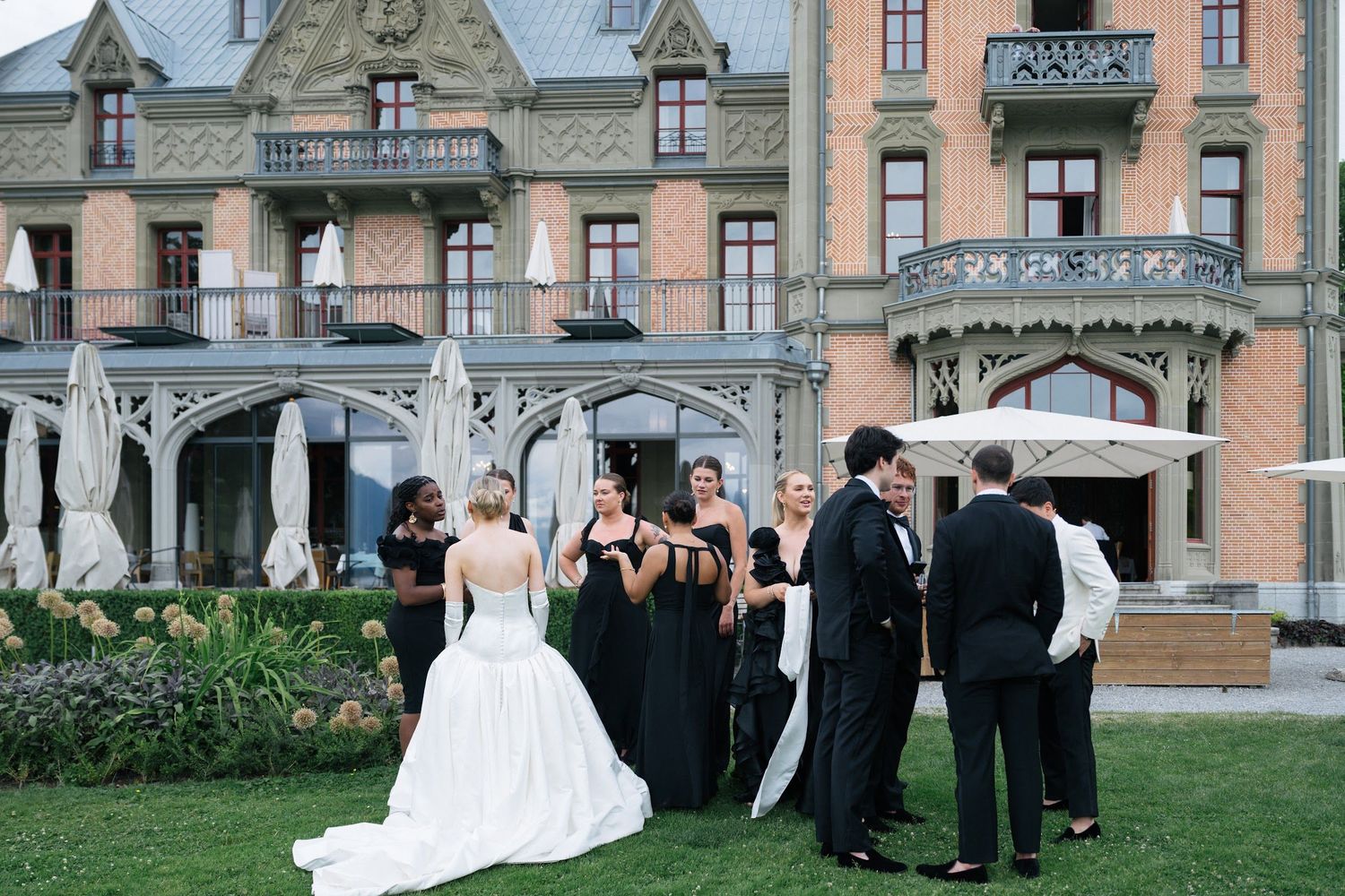Wedding party in black attire gathers outside ornate château venue with bride in white ball gown.