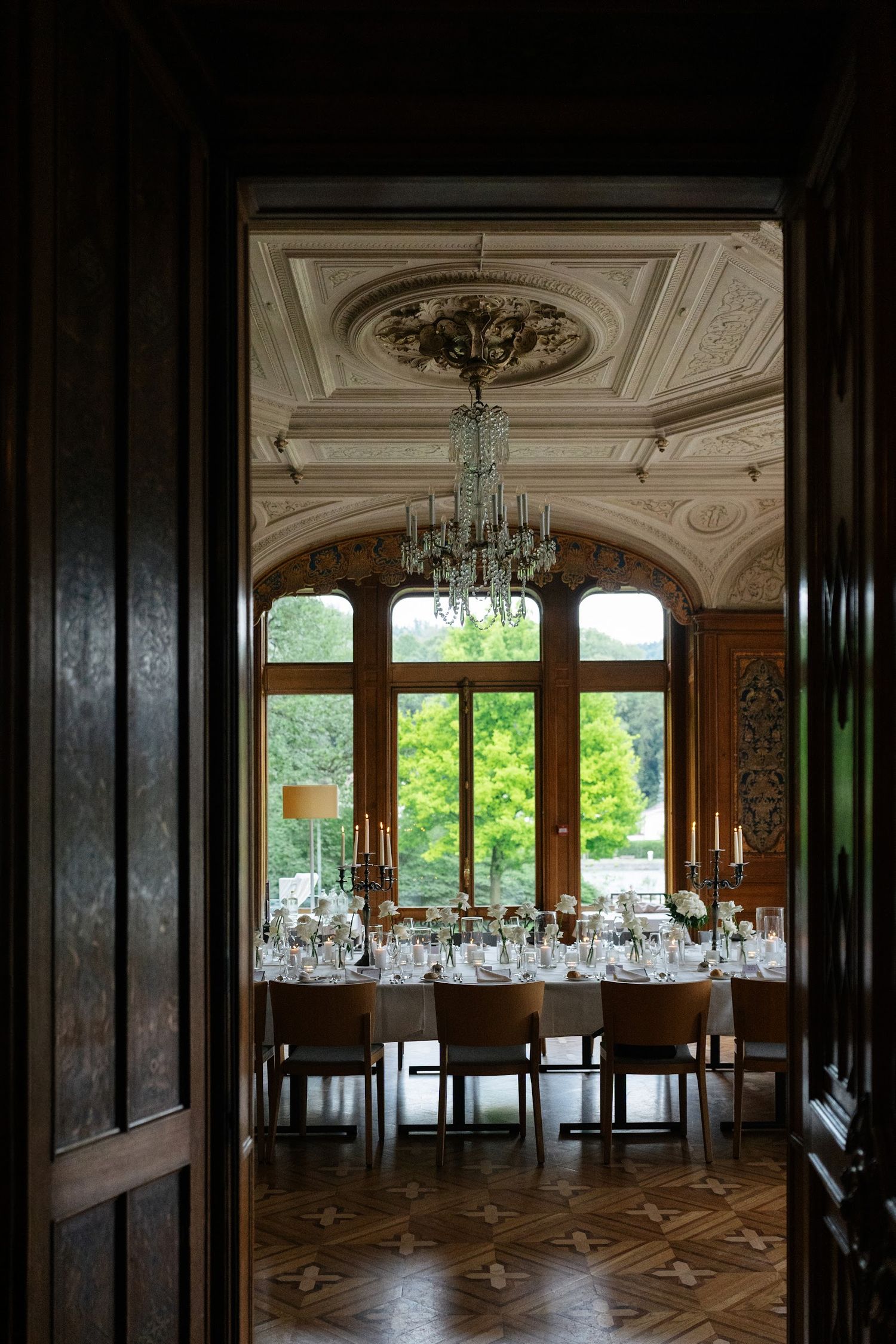 View through dark doorway into opulent château dining room with crystal chandelier and white wedding table.