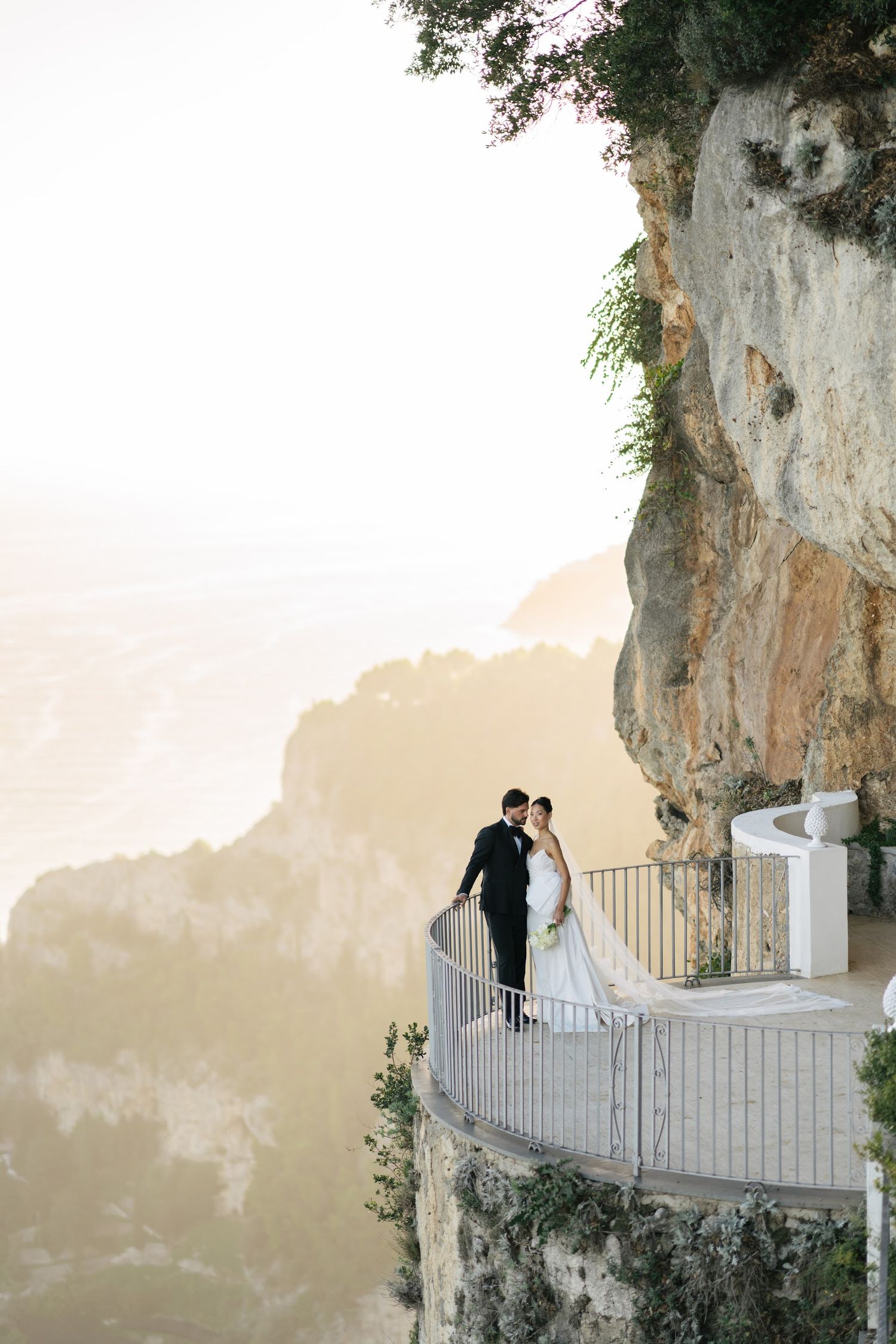Bride and groom embrace on a dramatic cliffside balcony overlooking the Amalfi Coast at golden hour.