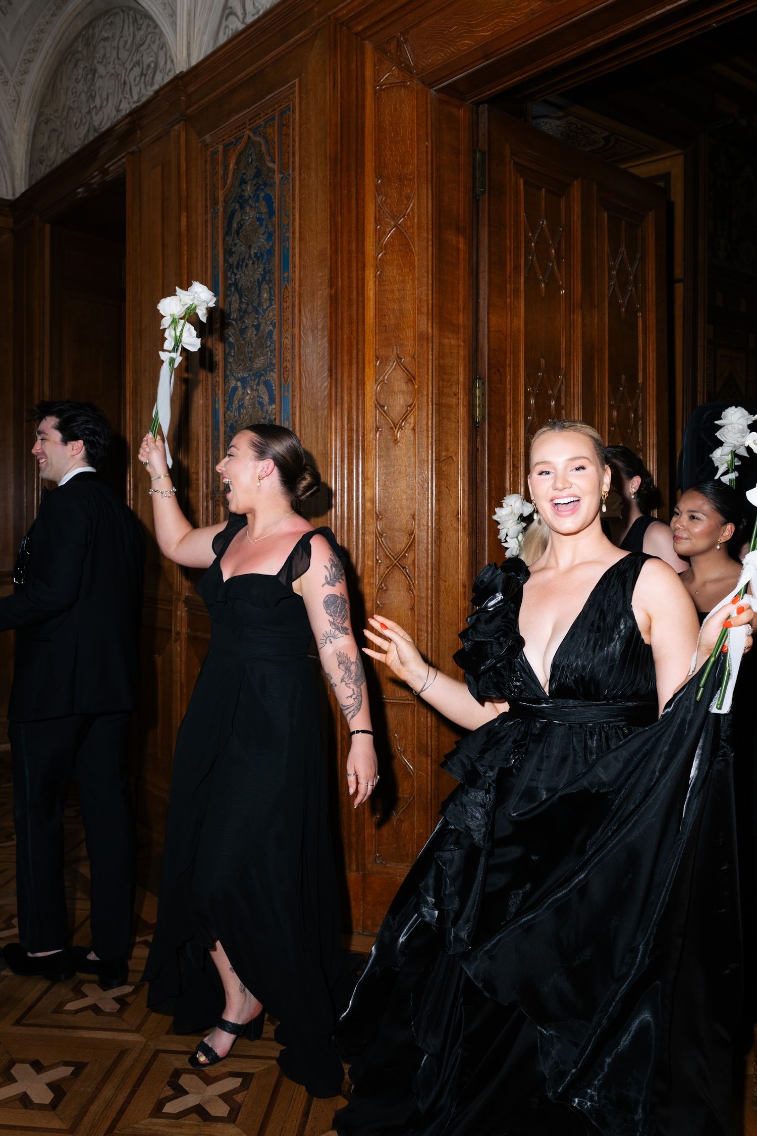 Bridesmaids in black gowns celebrate with white bouquets raised in an elegant wood-paneled venue.