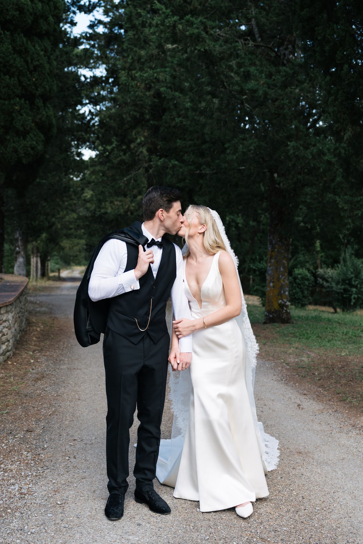 Groom kisses bride on a tree-lined gravel path after their elegant outdoor Tuscan wedding ceremony.