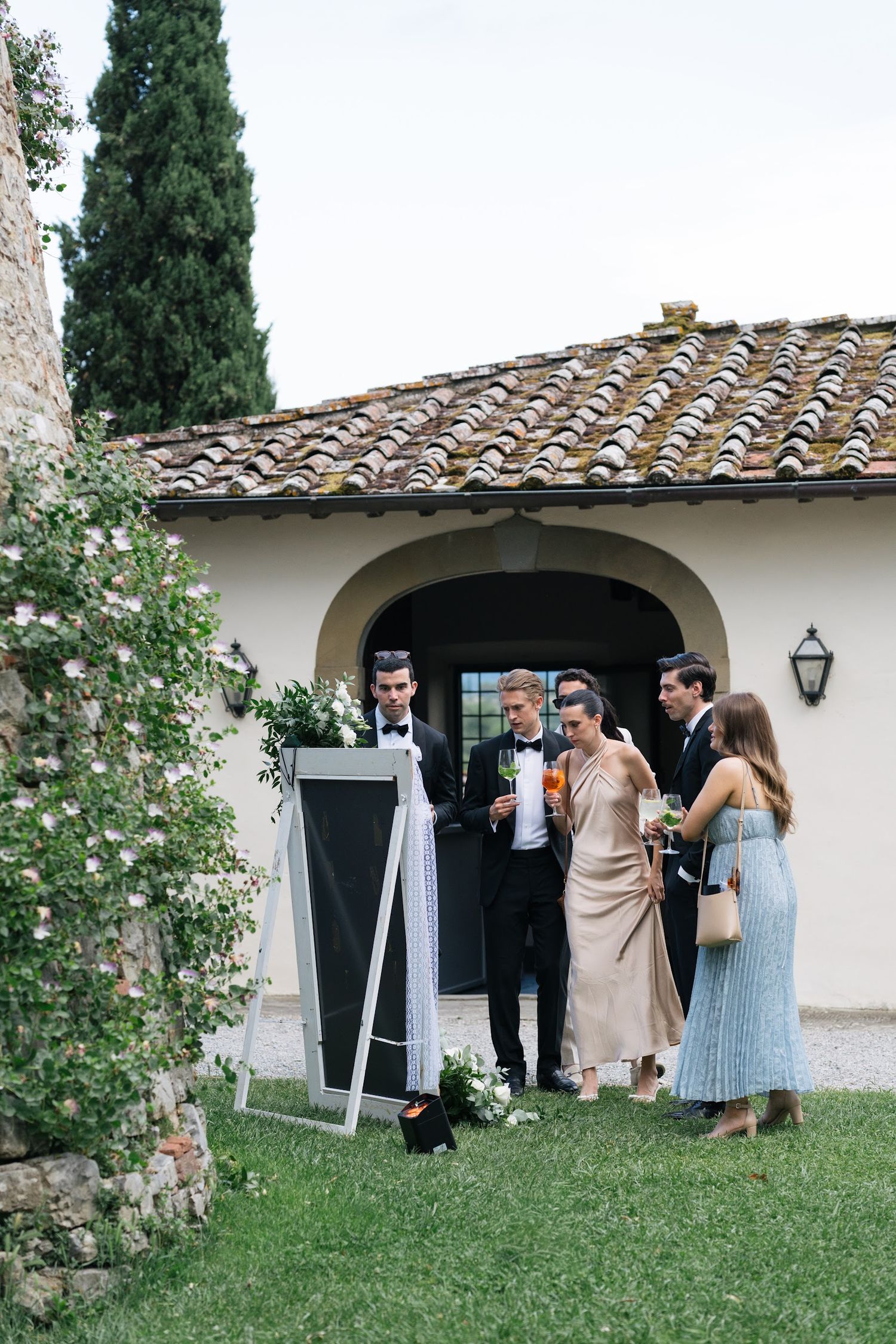 Wedding guests reviewing seating chart on an easel outside a Tuscan villa with terracotta roof.