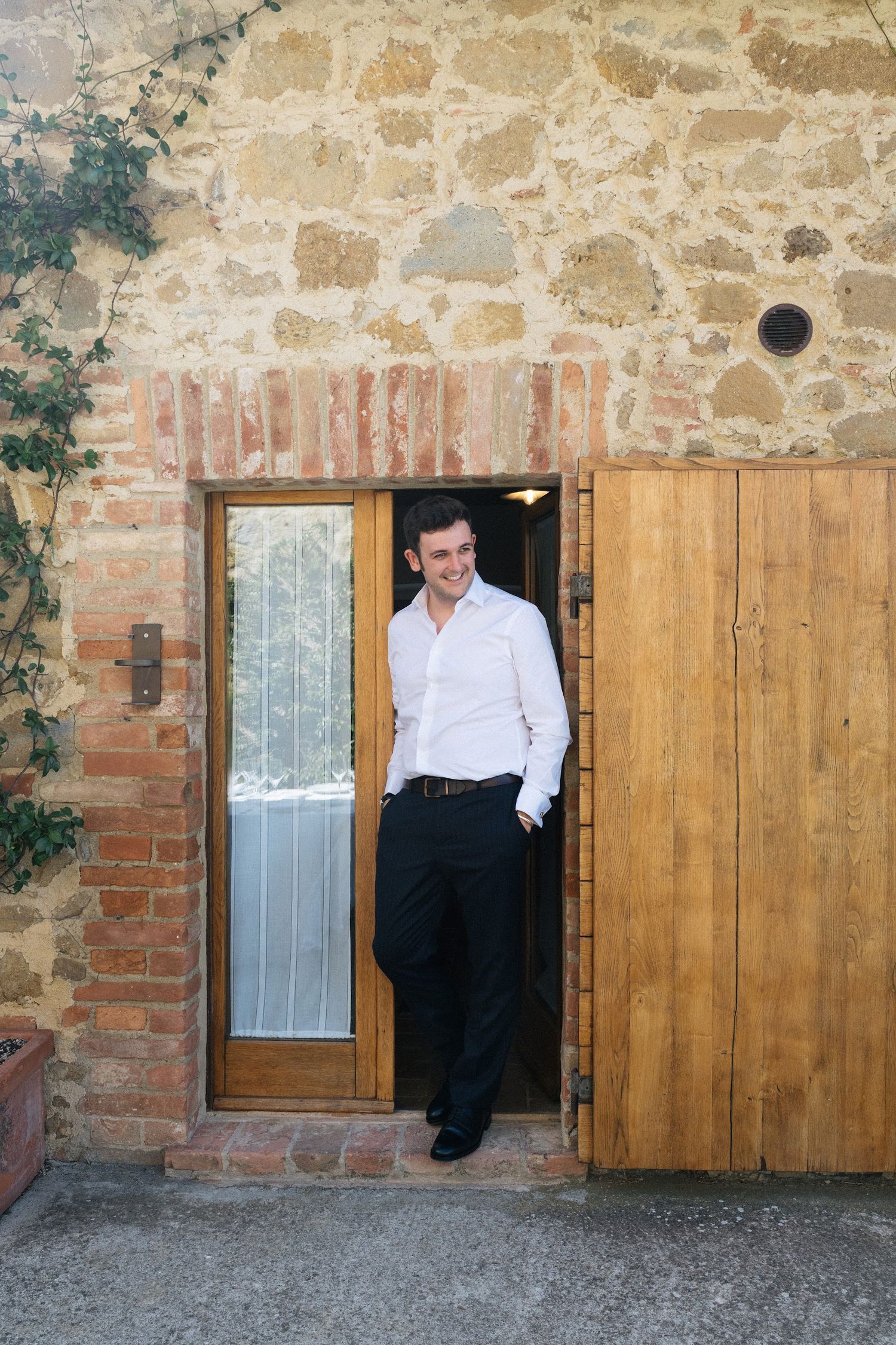 Smiling groom in white shirt and black trousers leaning against a wooden door of a rustic stone building.