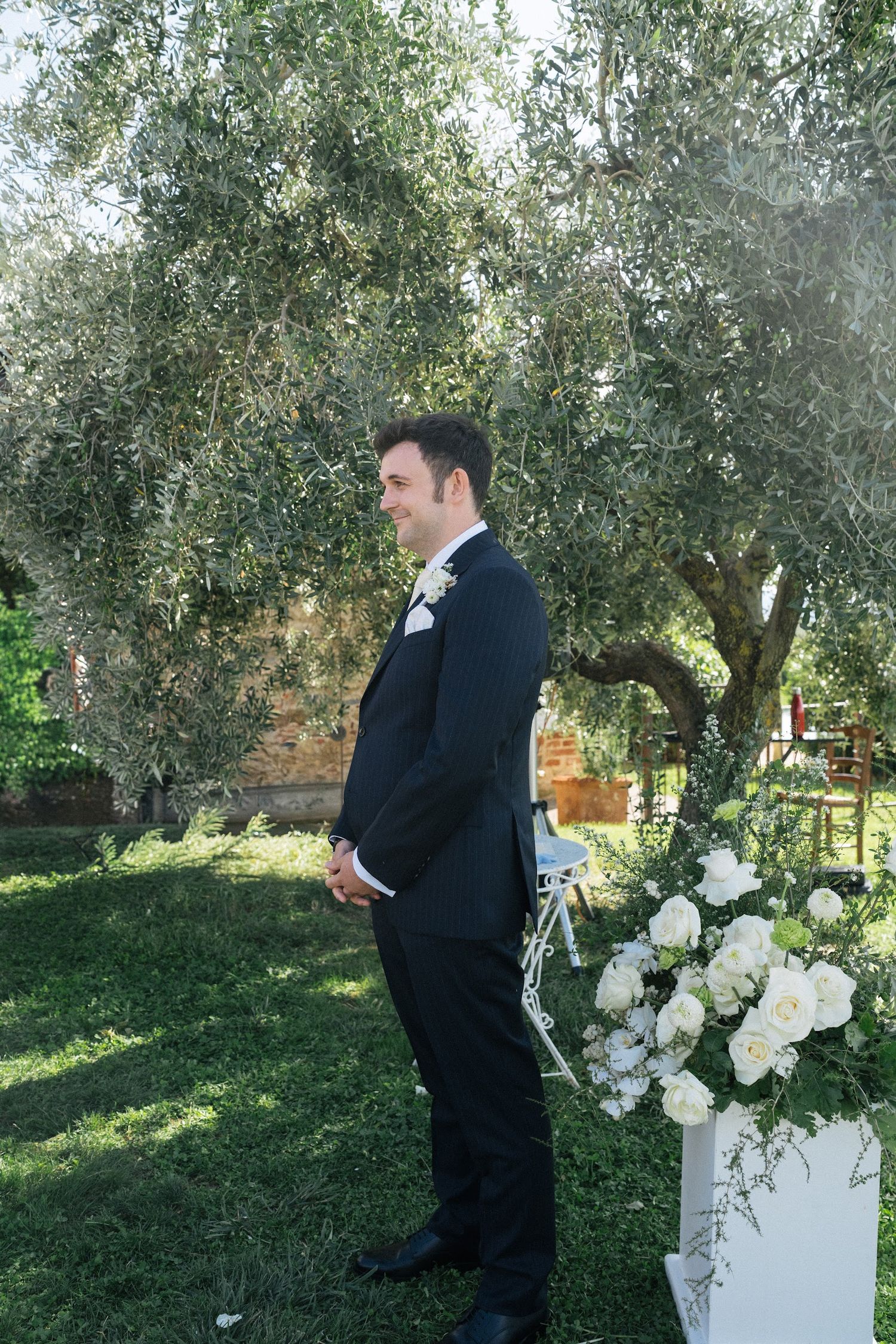 Smiling groom in black tuxedo standing under olive tree beside white floral arrangement at outdoor ceremony.