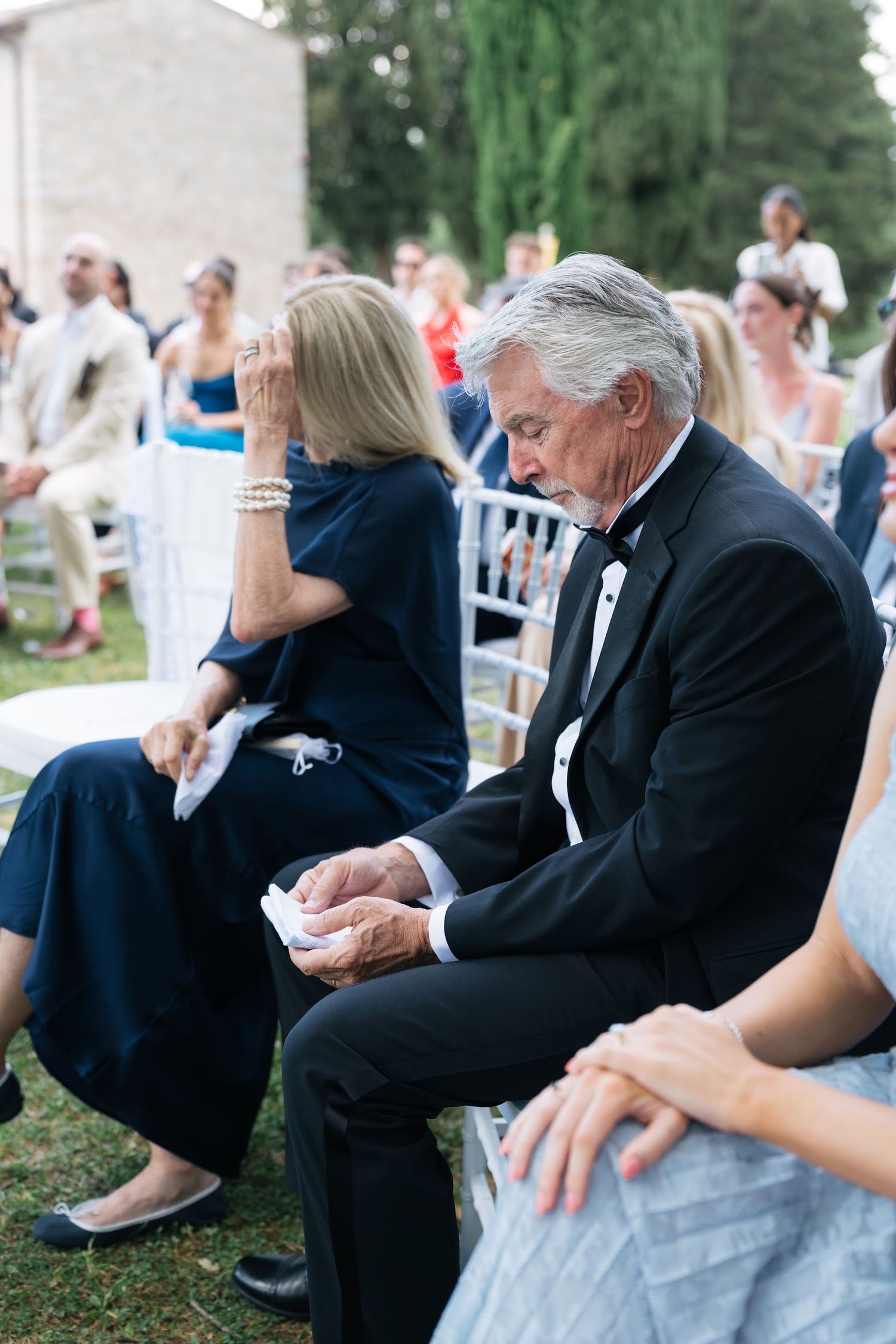 Emotional wedding guests in formal attire wipe tears during outdoor ceremony at Tuscan countryside venue.