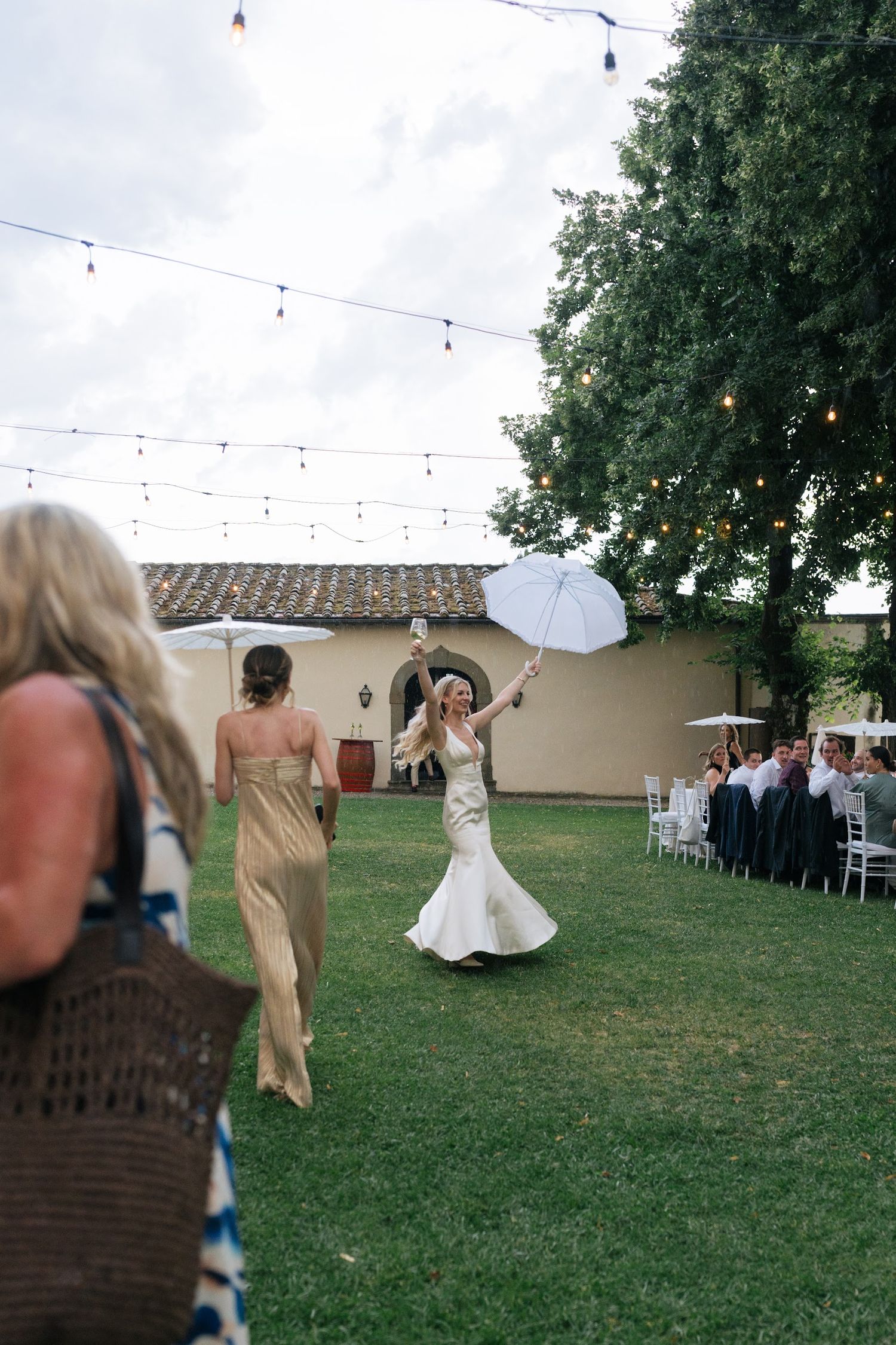 Bride twirling joyfully with white umbrella on lawn during outdoor wedding reception.