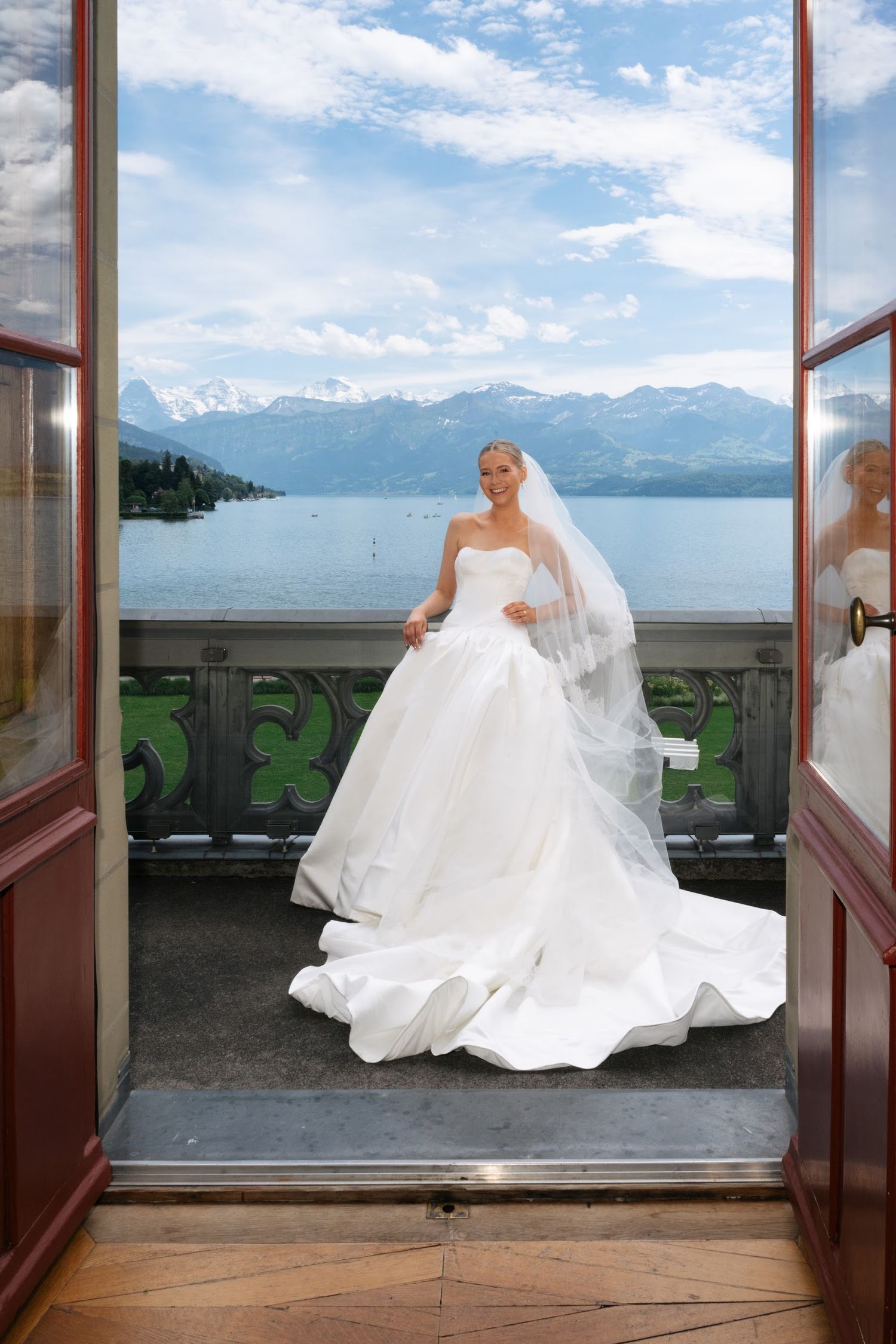 Radiant bride in white ball gown and veil posing on balcony with breathtaking lake and mountain backdrop.