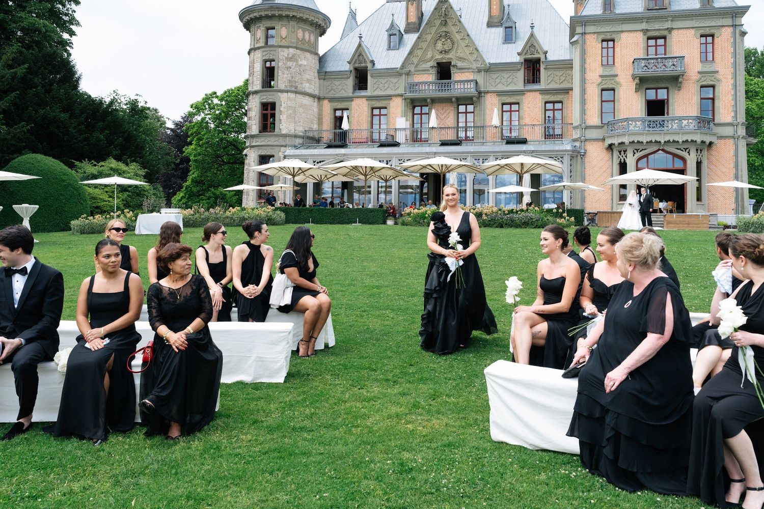 Wedding guests dressed in black gather on a lush lawn in front of a stunning historic château for an outdoor ceremony.