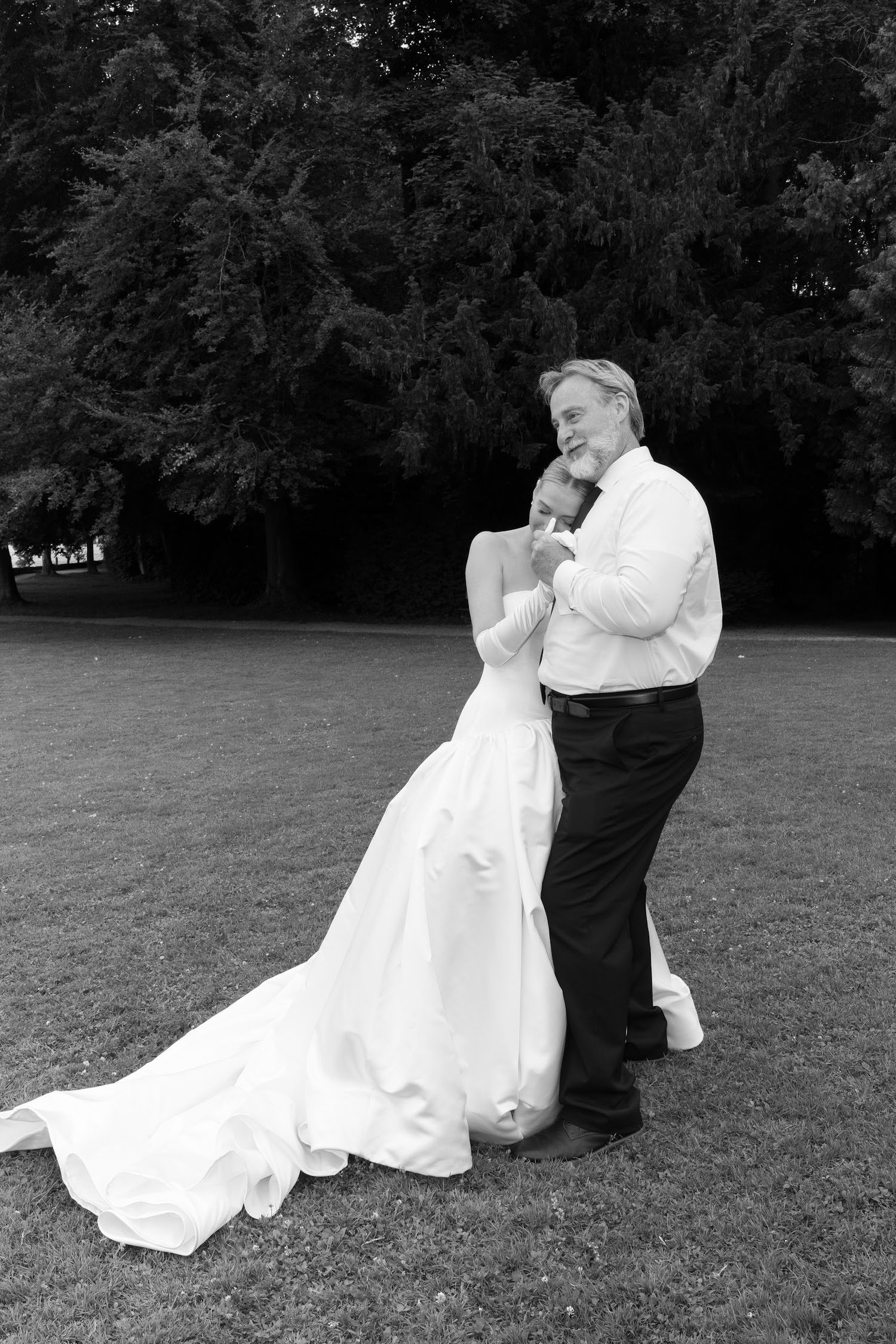 Black and white photo of bride in flowing gown dancing with older gentleman on garden lawn.