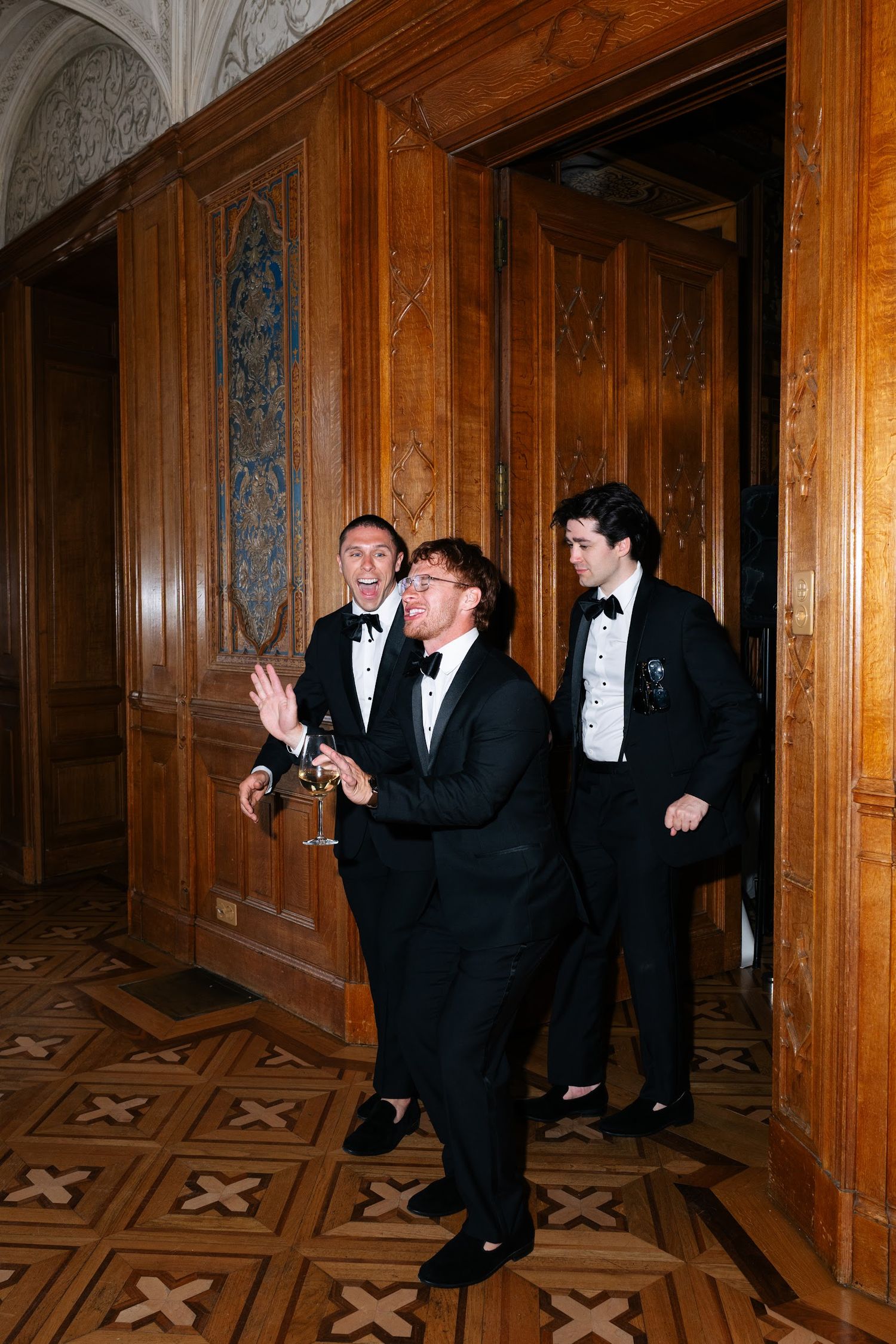 Three men in black tuxedos laughing and gesturing in an ornate wood-paneled château doorway.