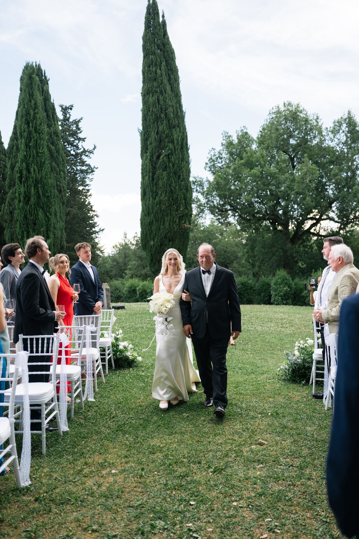 Bride walks down grassy aisle escorted by father at elegant outdoor Tuscan wedding with cypress trees.