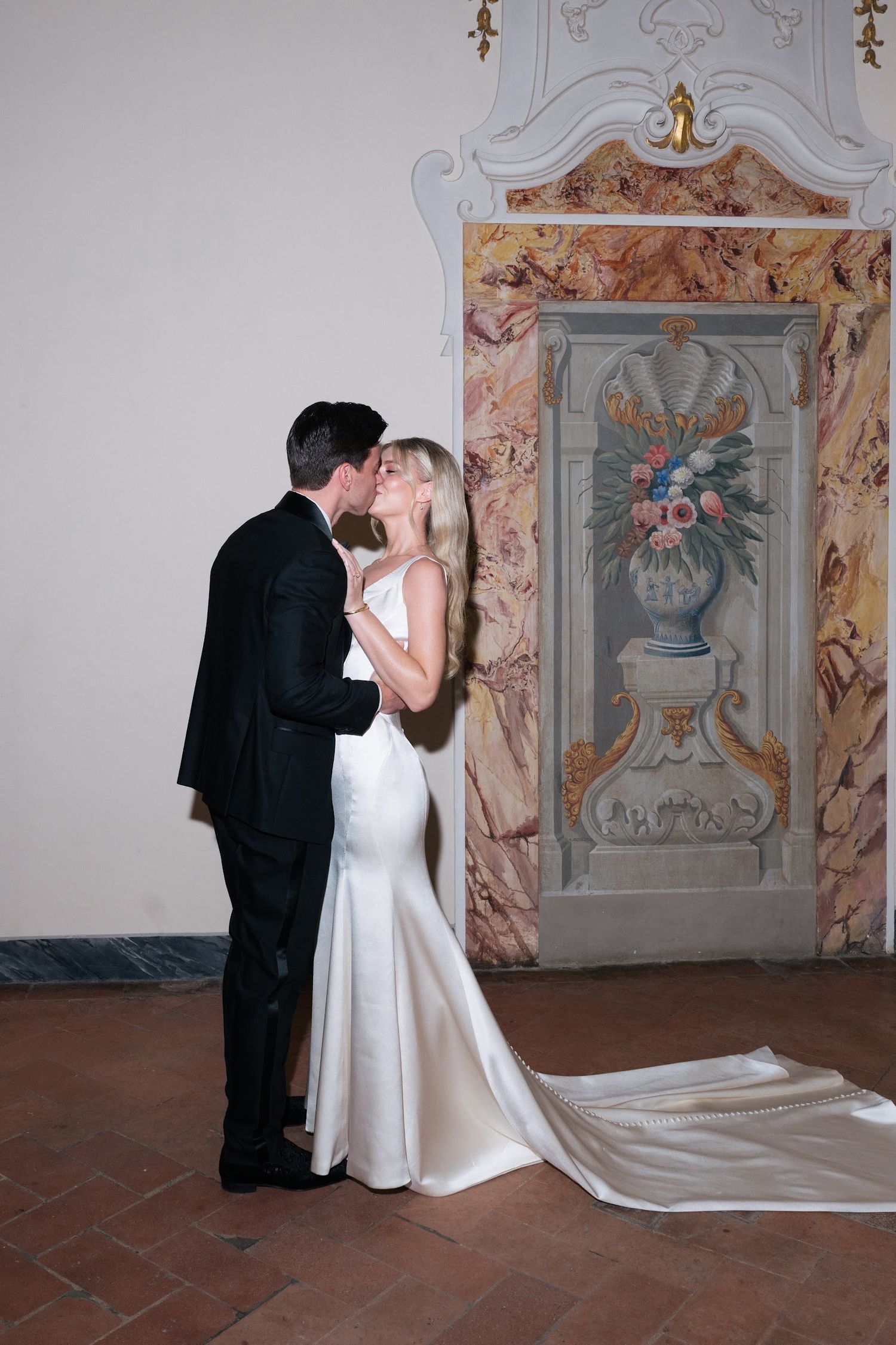 A bride and groom share a romantic kiss in front of an ornate floral fresco at their wedding venue.