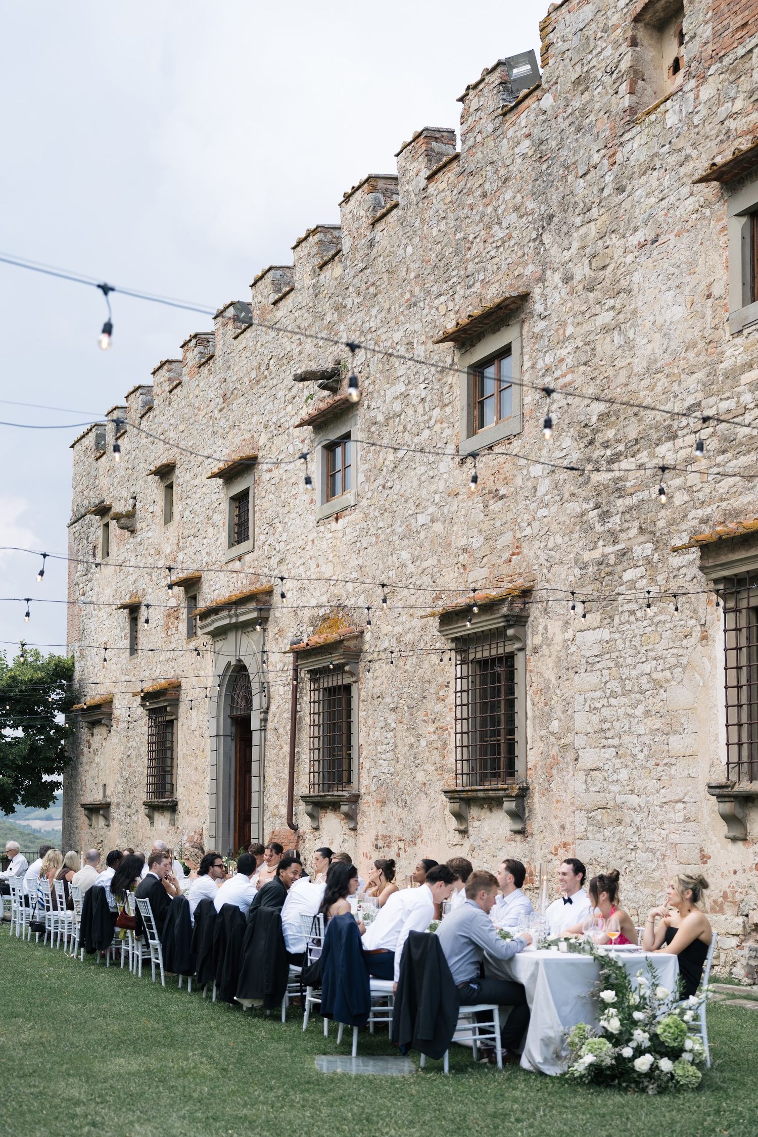 Elegant outdoor wedding reception with long tables set along a historic Italian stone castle facade.