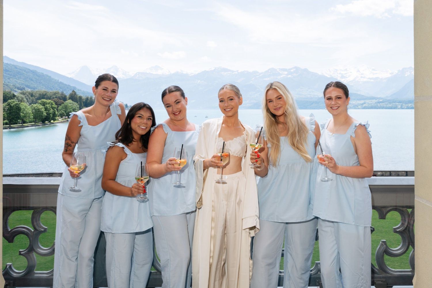 Bride and bridesmaids in matching light blue outfits toasting champagne on a balcony with lake views.