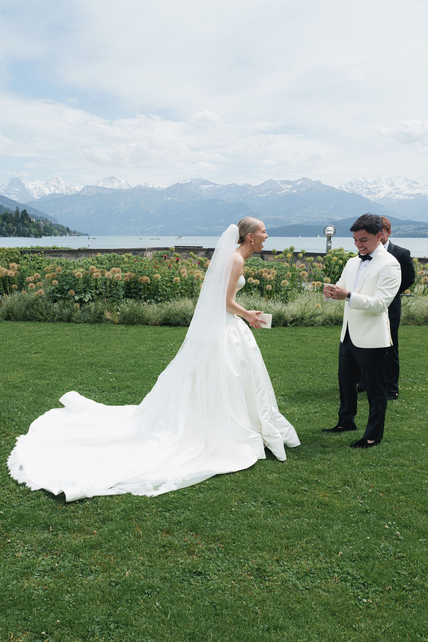 Bride in white gown with long train and groom in white tuxedo jacket exchange vows outdoors with mountain lake backdrop.