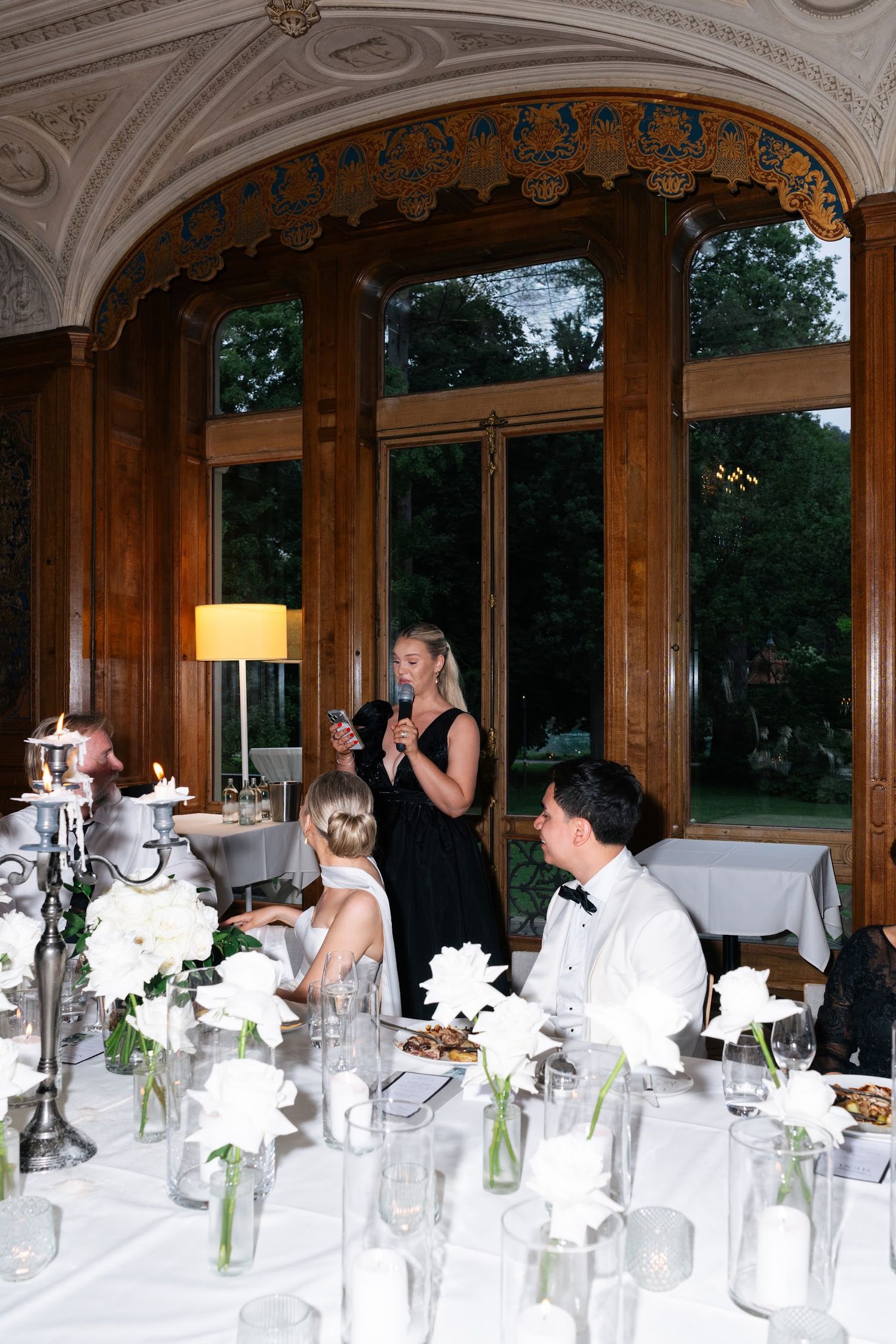 Wedding guest gives a speech at reception while bride and groom sit at elegantly decorated table.