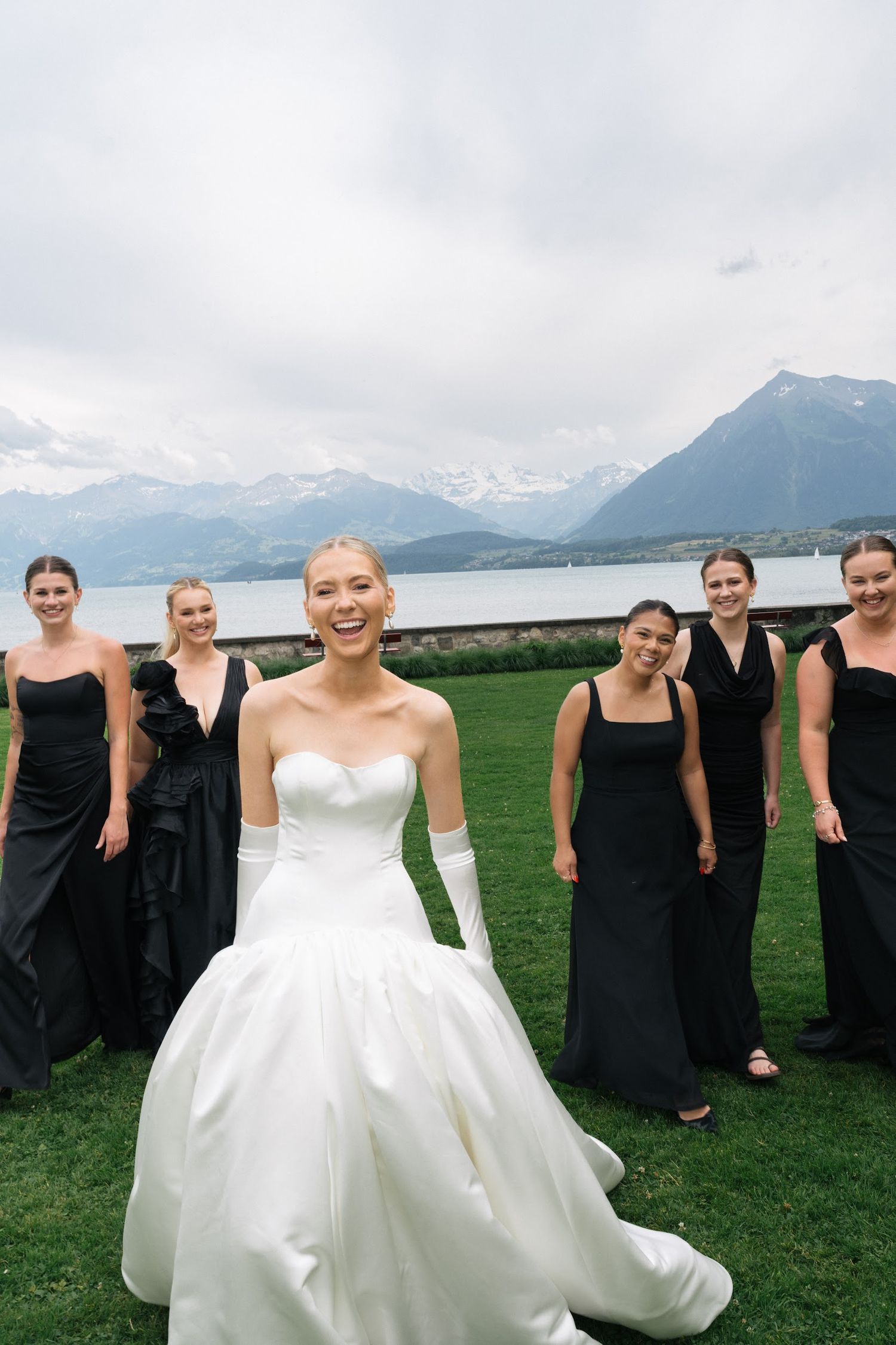 Joyful bride in strapless white gown laughing with bridesmaids in black dresses by alpine lake.