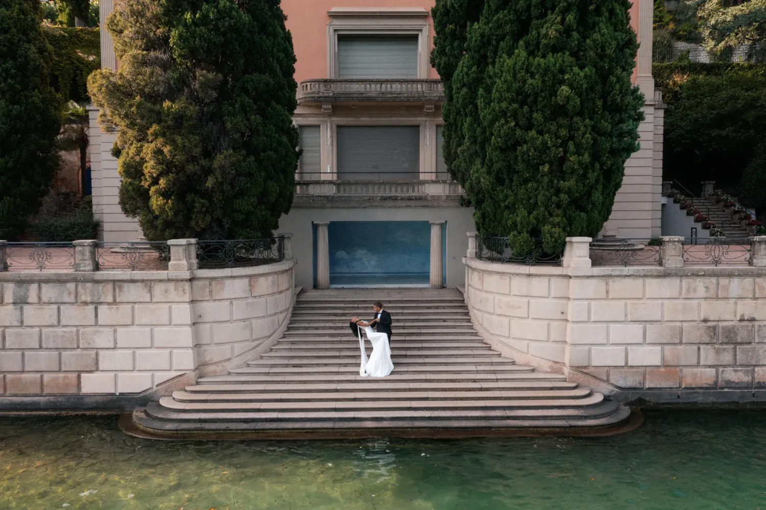 Bride and groom pose romantically on stone steps beside a Villa in Lugano, Ticino surrounded by cypress trees.