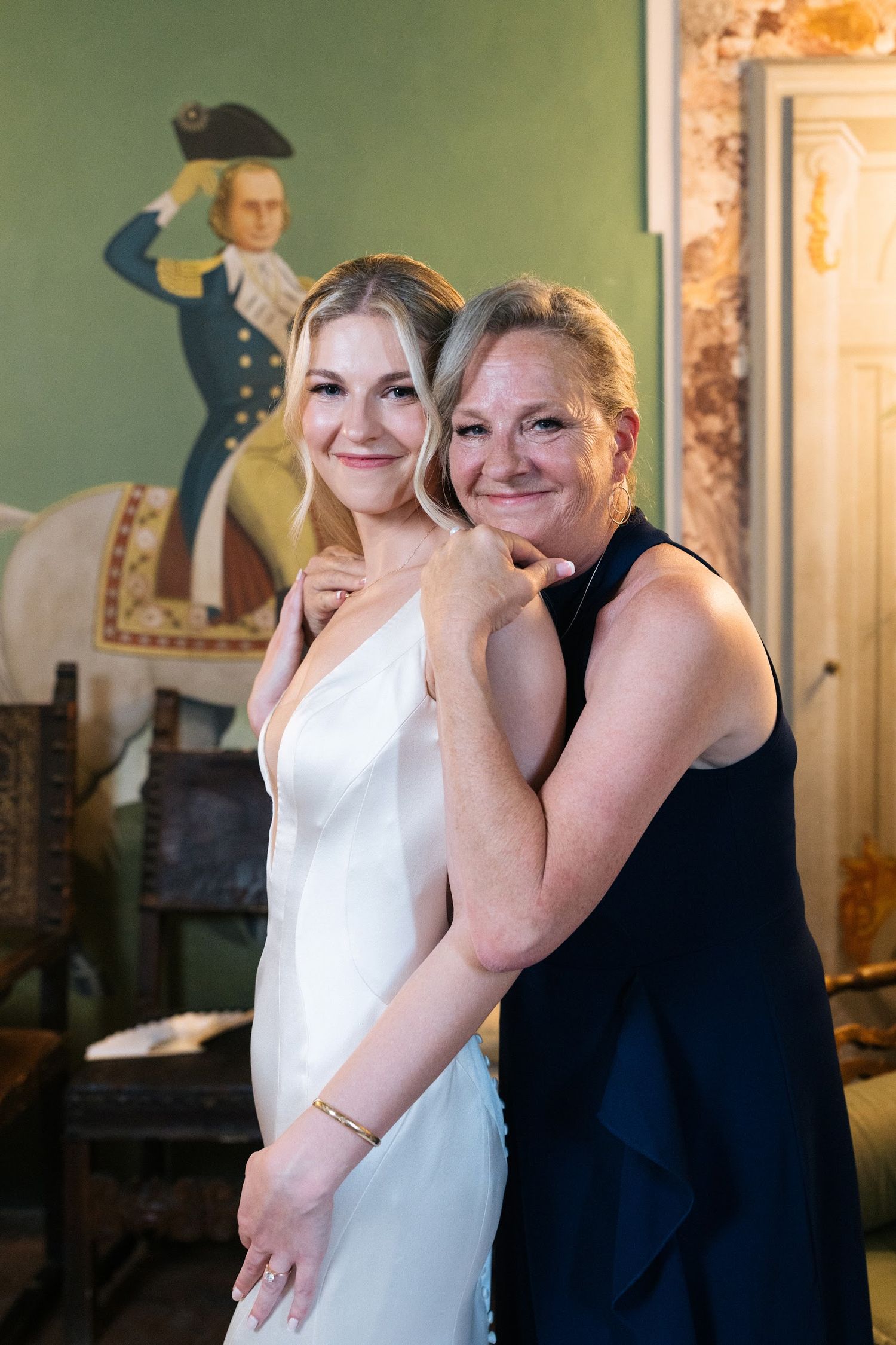 Smiling bride in white satin gown embracing her mother in front of a historic portrait painting.