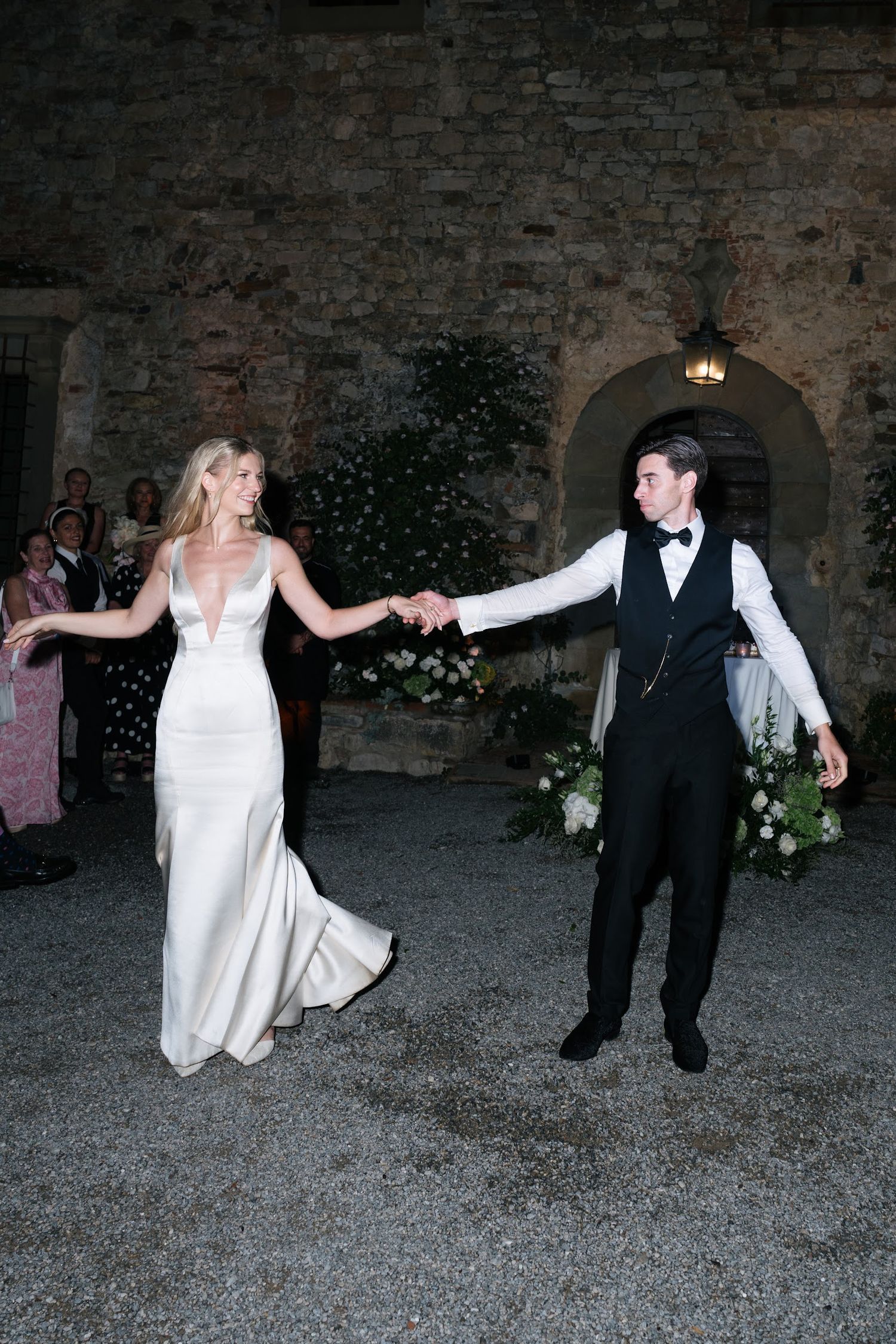 Bride in white gown and groom in black tuxedo holding hands during outdoor first dance at night.