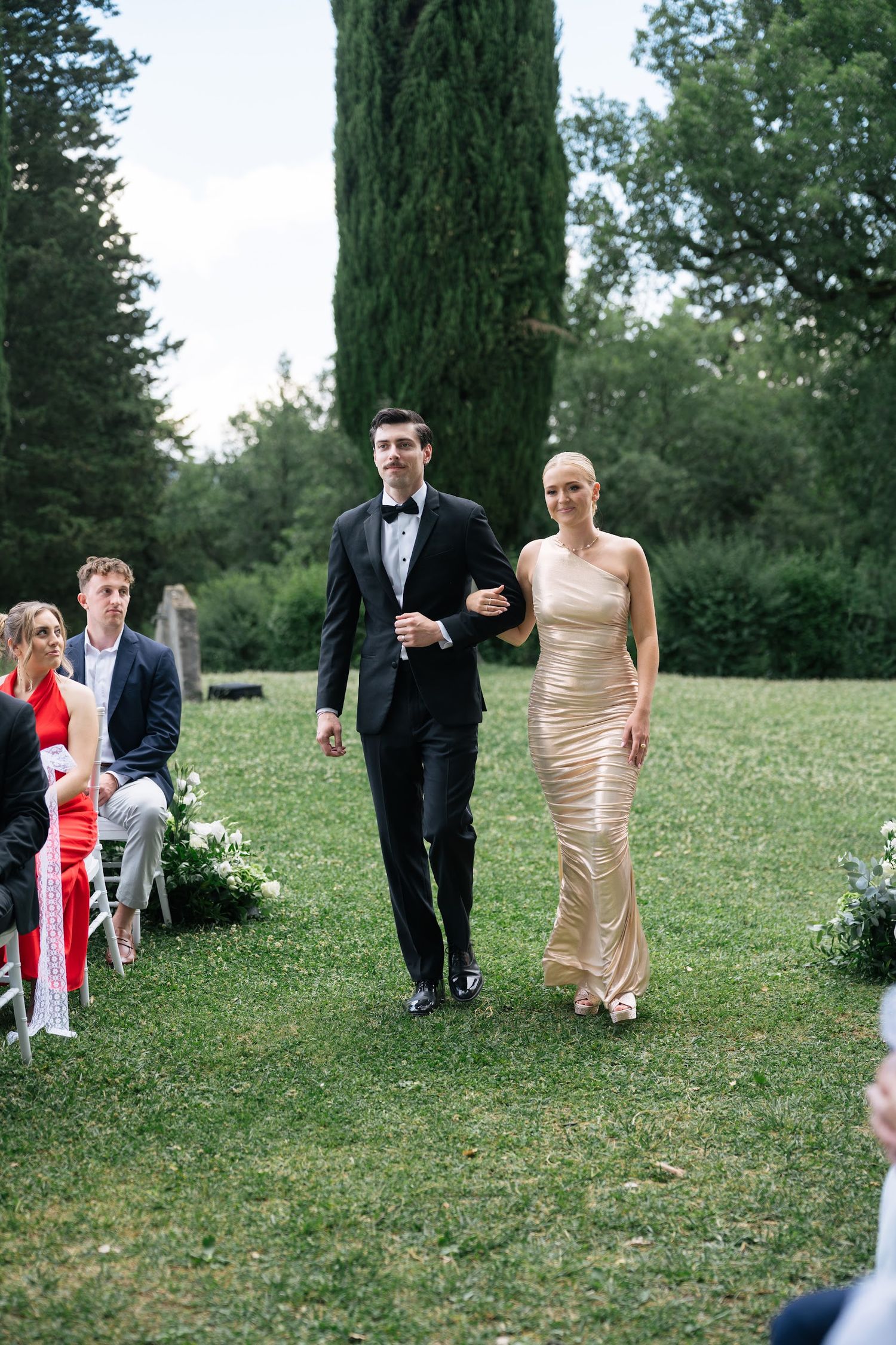 Groomsman in black tuxedo escorts bridesmaid in gold gown down aisle at outdoor Tuscan wedding ceremony.