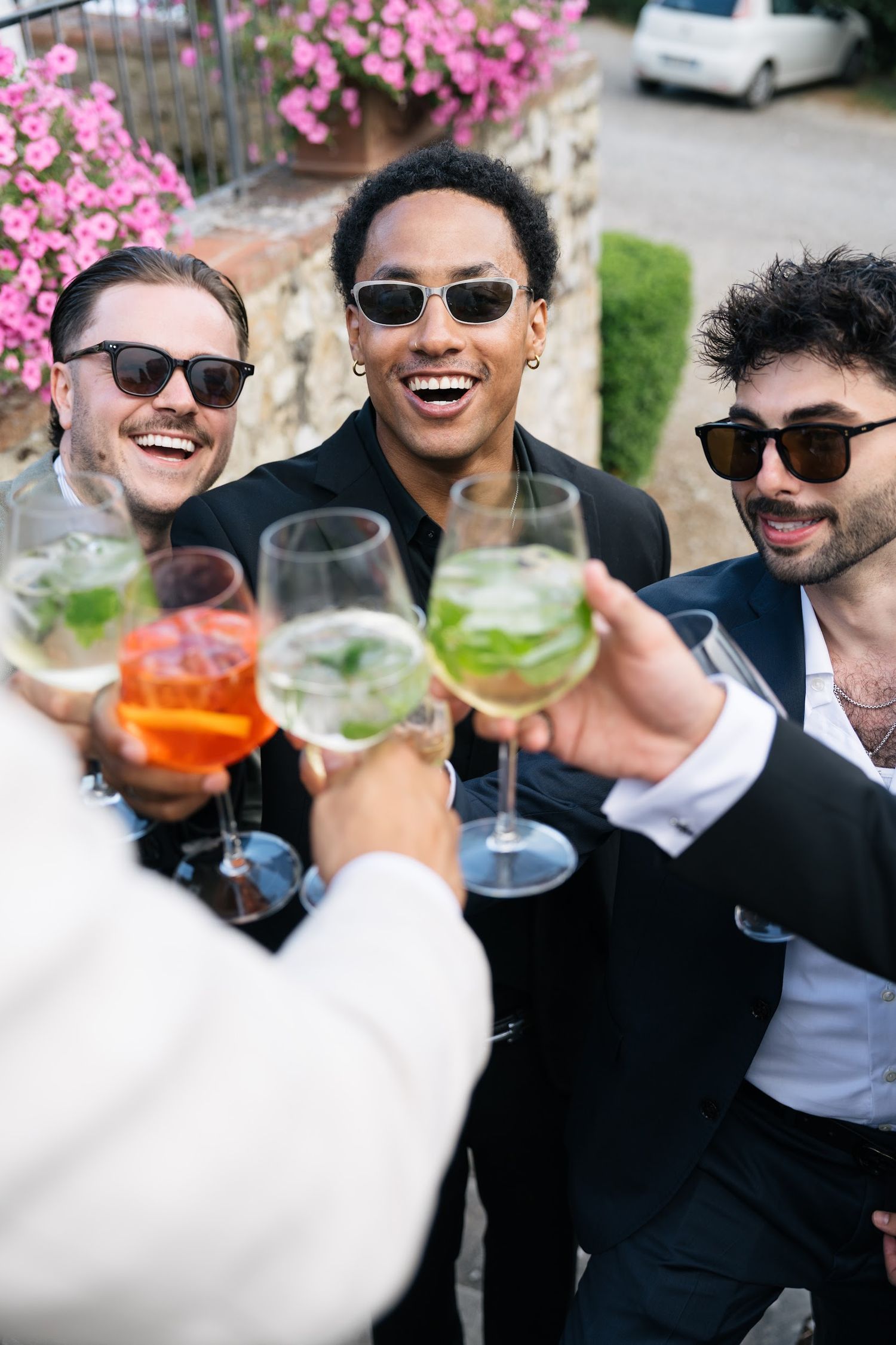 Three groomsmen in black suits and sunglasses toasting with colorful cocktails at an outdoor wedding.