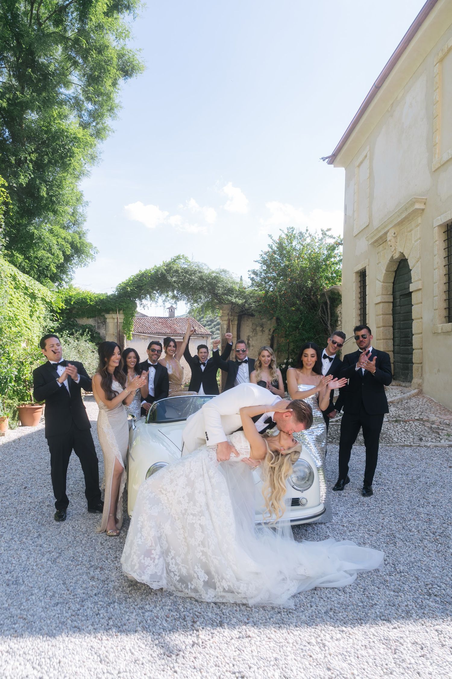 Groom dips bride near vintage car as wedding party celebrates at Italian villa, Verona.