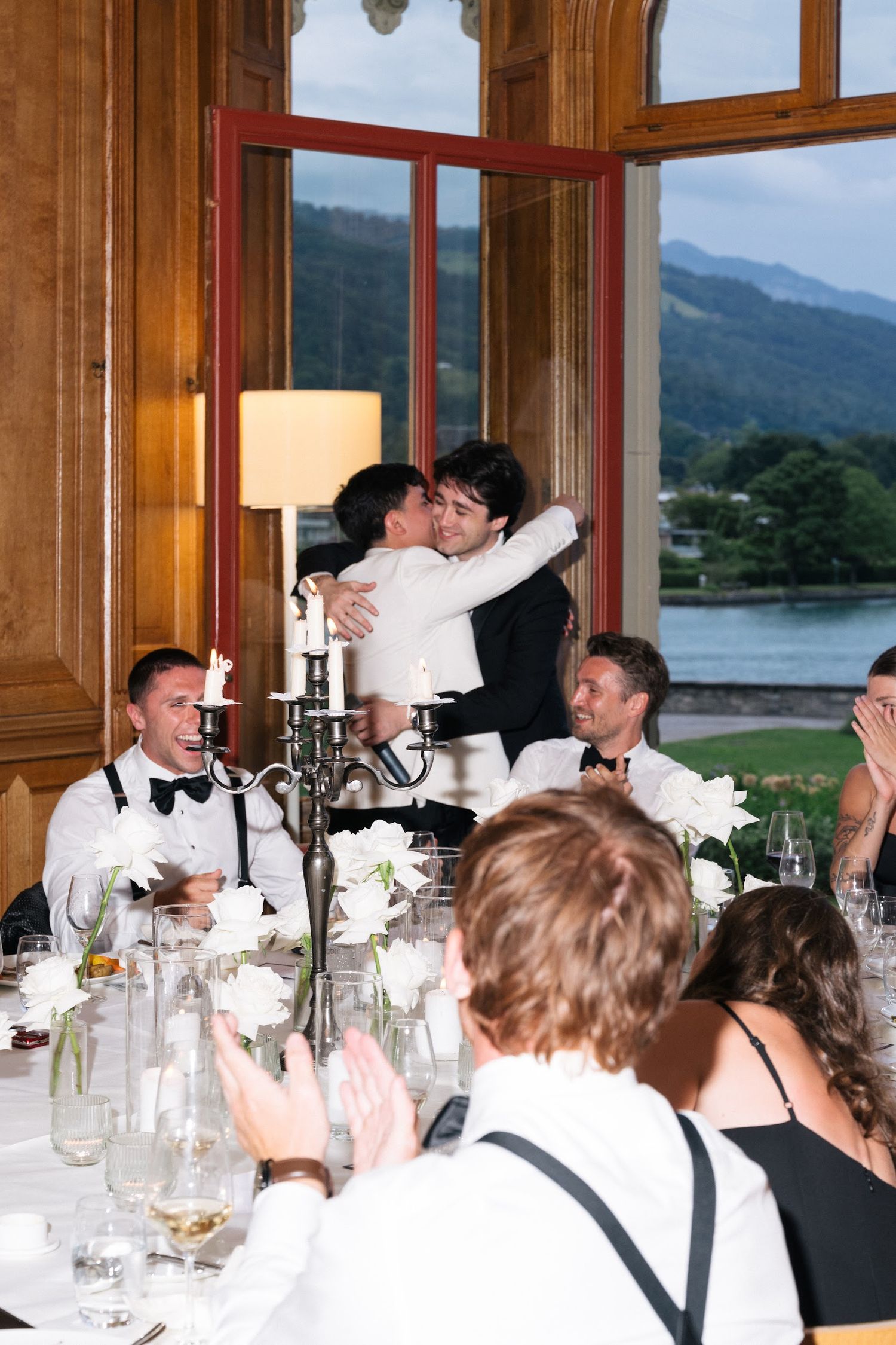 Wedding guests embracing and celebrating at a lakeside reception dinner with white floral decor.