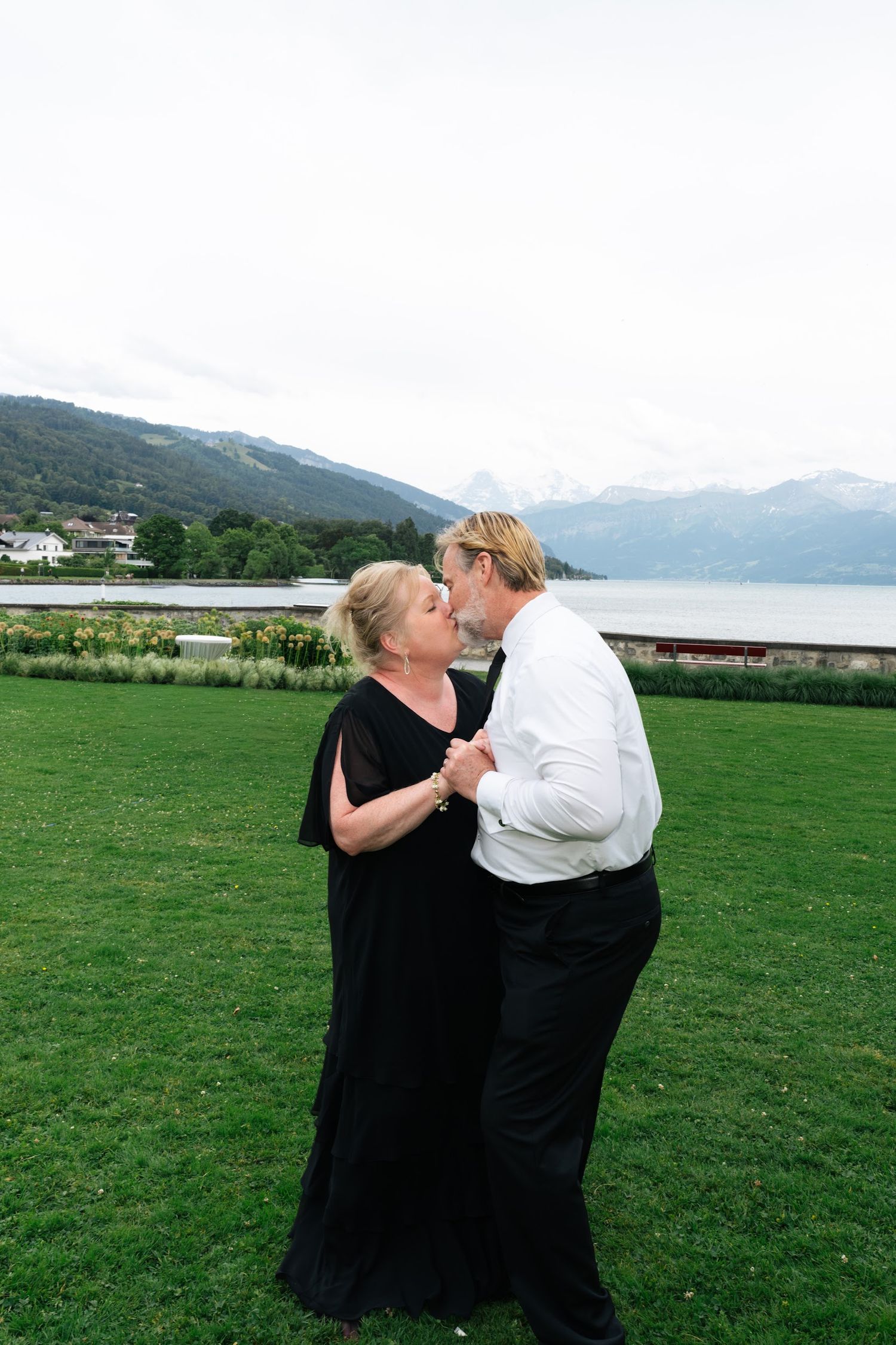 Older couple sharing a romantic kiss on green lawn with alpine lake and mountain scenery behind.