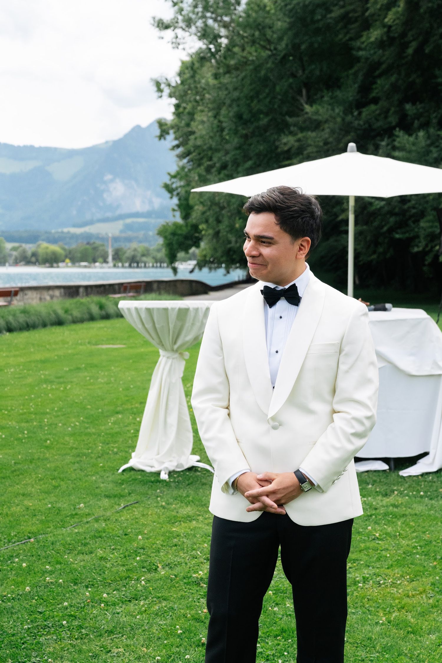 Dapper groom in a white tuxedo jacket and black bow tie stands on a green lawn with mountain lake views behind him.