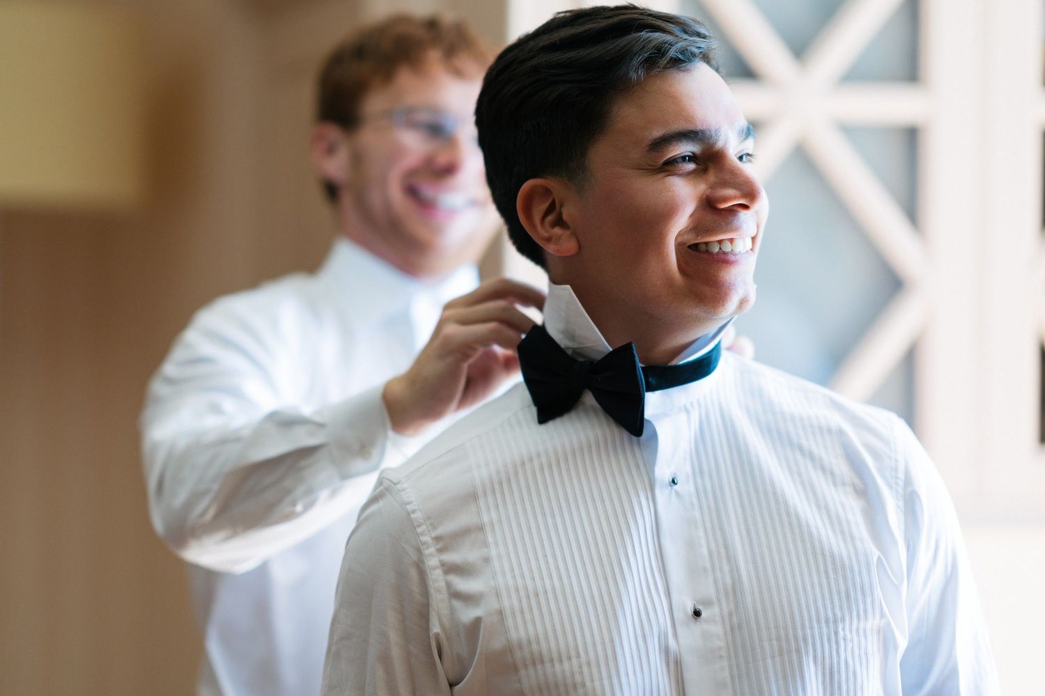 Two groomsmen in tuxedo shirts smiling as one adjusts the other's black bow tie before wedding ceremony.