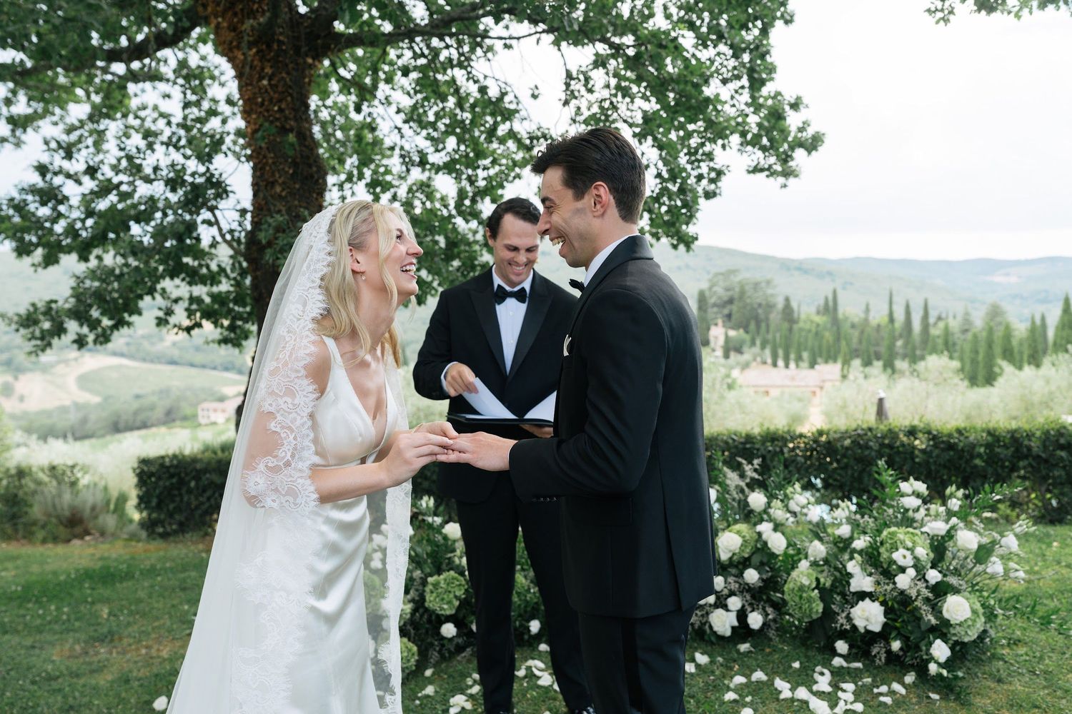 Bride and groom exchange vows outdoors in Tuscany with rolling hills backdrop and white floral arrangements.