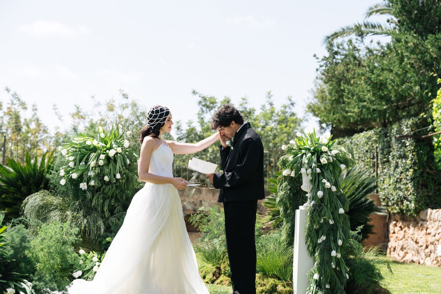 Bride and groom exchanging vows in a lush outdoor garden ceremony with white floral arches.