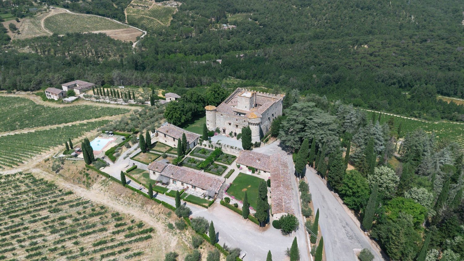 Aerial view of historic Tuscan castle surrounded by manicured gardens, cypress trees, and sprawling vineyards.