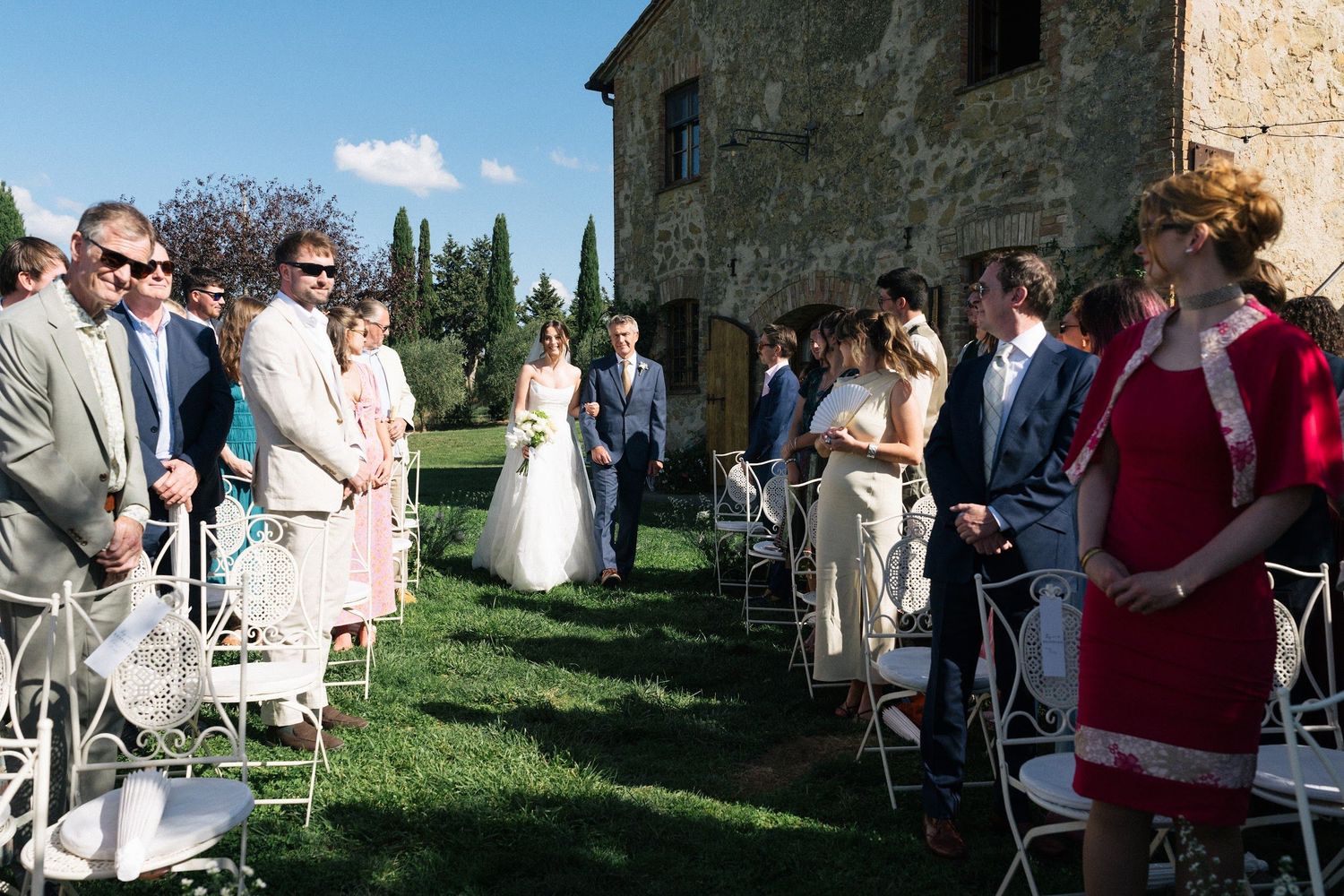 Wedding guests standing as bride walks down outdoor aisle at rustic Tuscan stone villa ceremony.