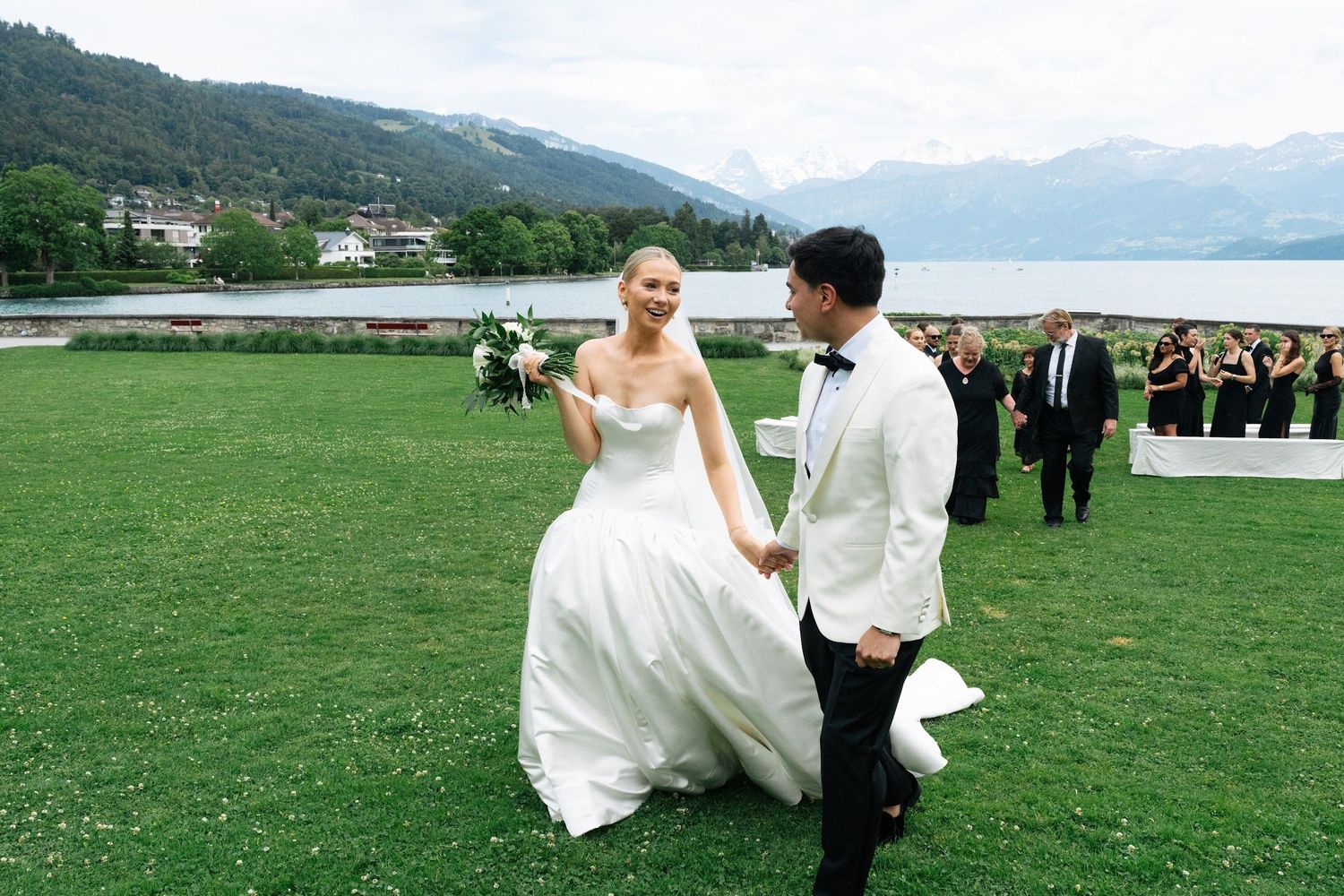 Smiling newlyweds walk hand in hand across a green lawn with an alpine lake and mountains in the background.