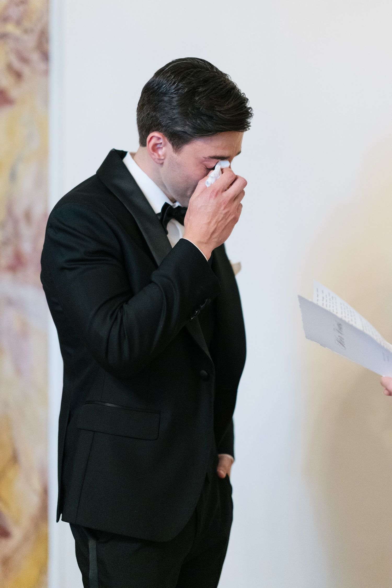 An emotional groom in a black tuxedo dabs his eyes with a handkerchief during private vow reading.