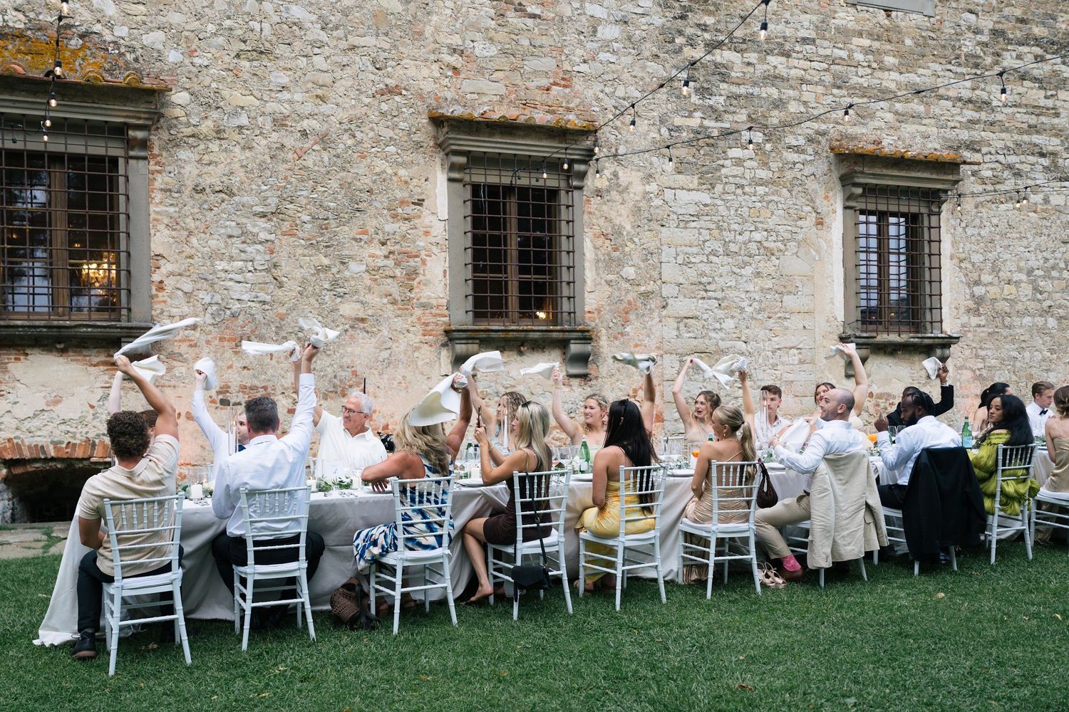 Wedding guests wave napkins in celebration at an outdoor reception beside a rustic Italian stone castle venue.