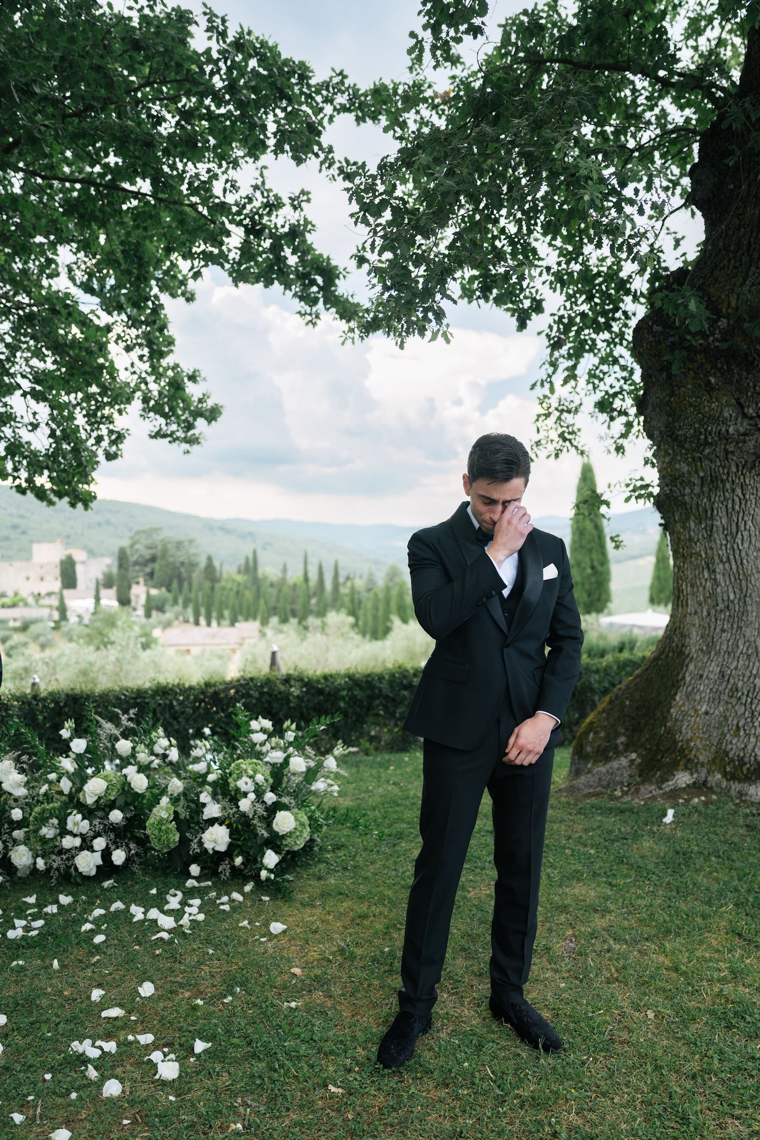 Emotional groom in black tuxedo wipes tear beside white floral arch with Tuscan landscape behind him.