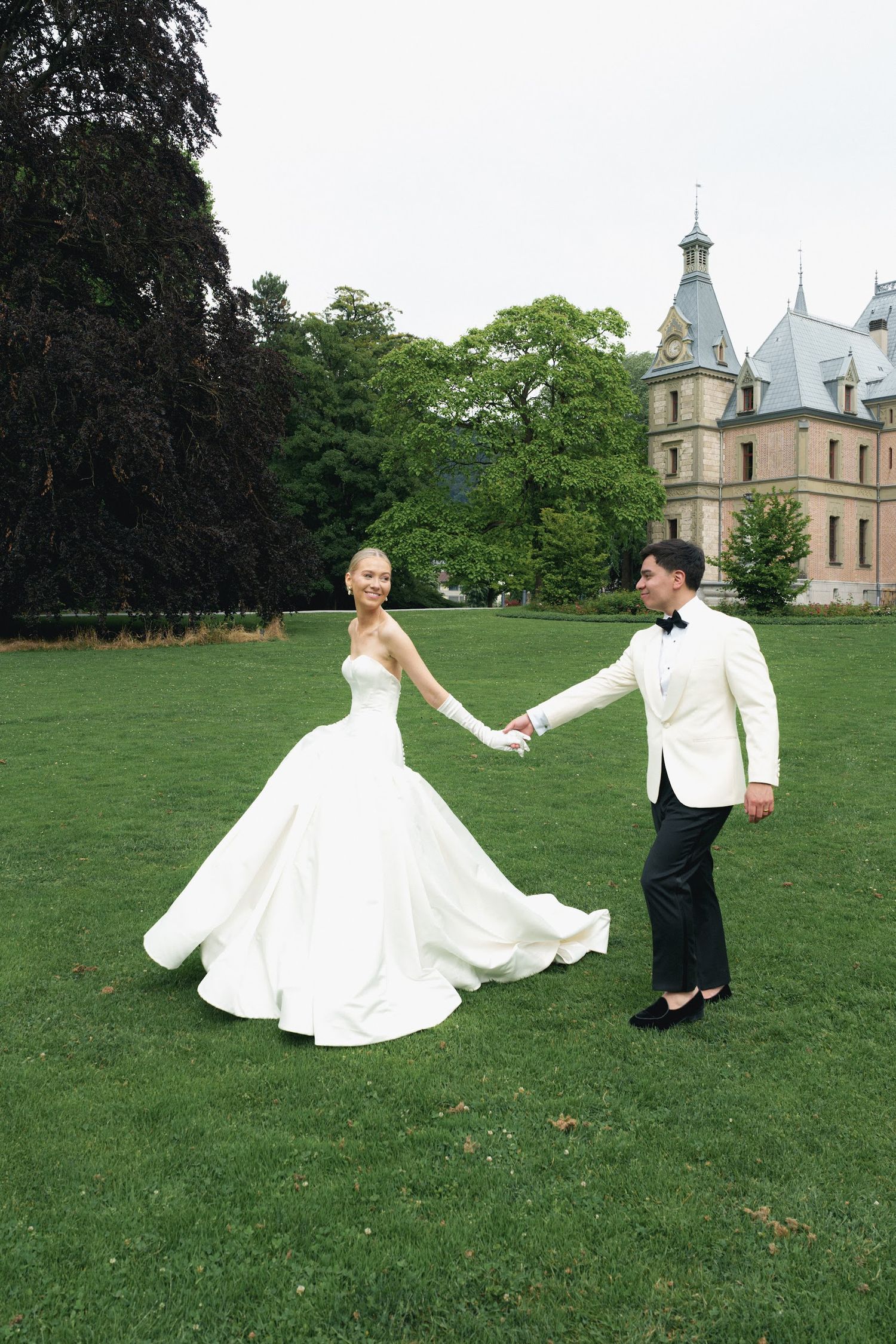 Bride in voluminous white ballgown twirls with groom in white tuxedo on lawn of grand castle estate.